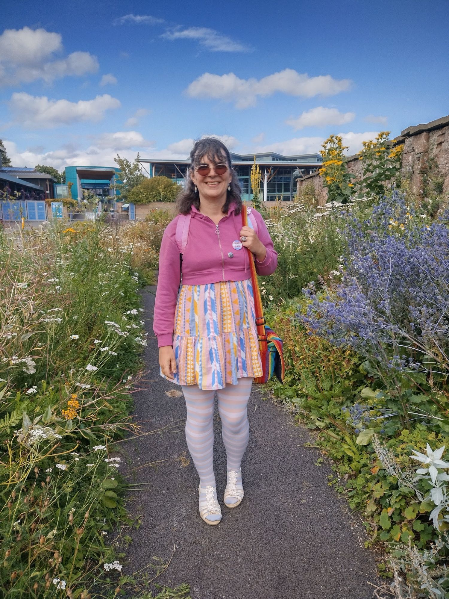 Me standing on a footpath with wild flowers growing either side and some modern style glass steel buildings behind me. I am wearing a pink crop hoodie over a pastel dress, with trans flag striped tights and white sandals. I have a rainbow handbag. Shades, dangly earrings and a couple of pronoun pins complete my look.