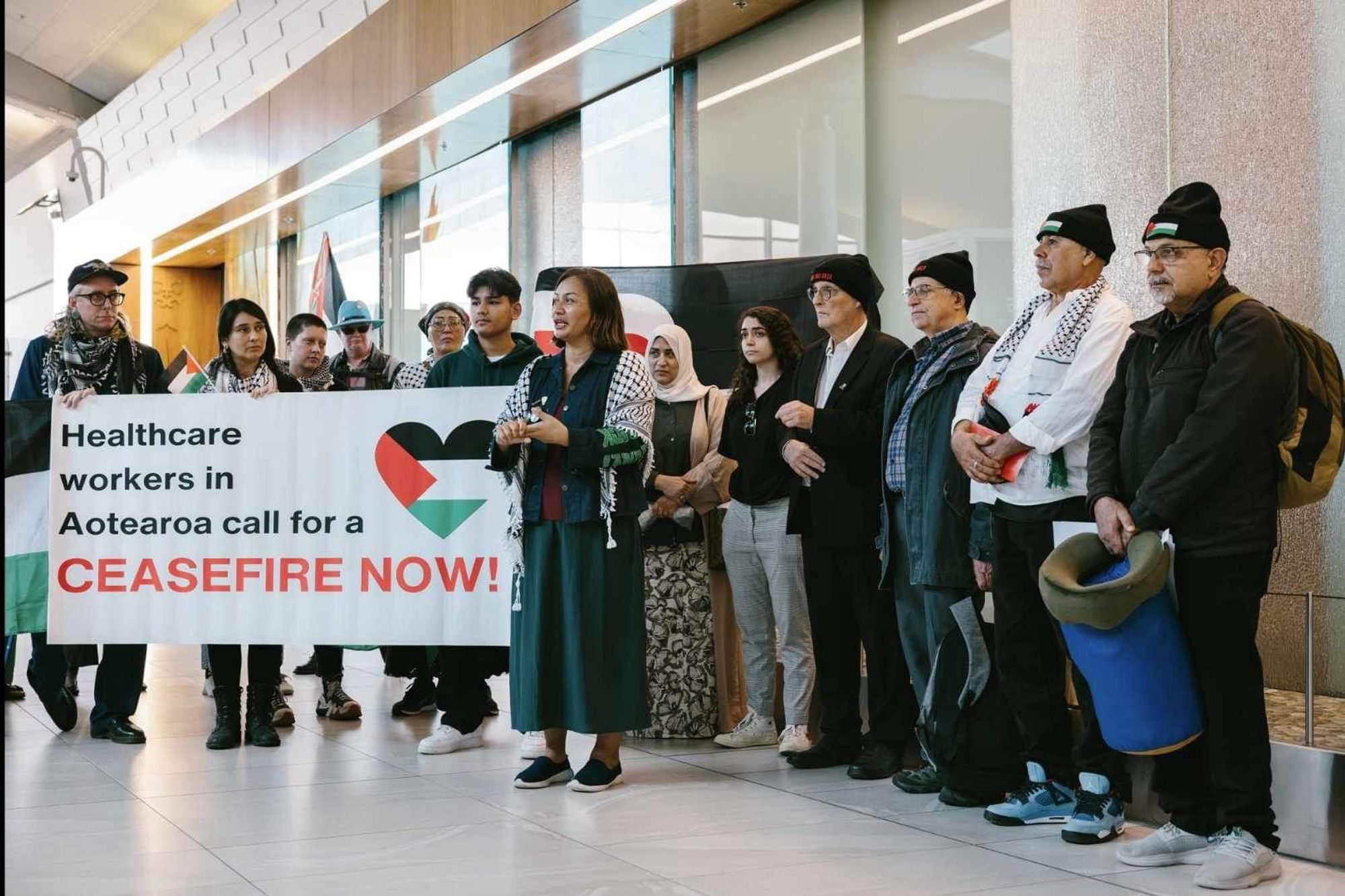 Three Kiwi doctors and supporters, holding a banner reading “Healthcare Workers in Aotearoa call for a CEASEFIRE NOW!”, at Auckland International Airport