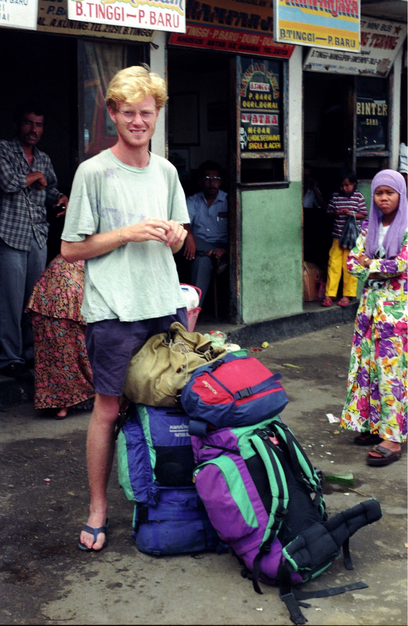 A skinny young man (me, a long time ago) stands guard over two rucksacks in a bus station. Behind him is a sign for the bus from Bukittinggi to Pekanbaru. Some locals stand around looking at the camera.