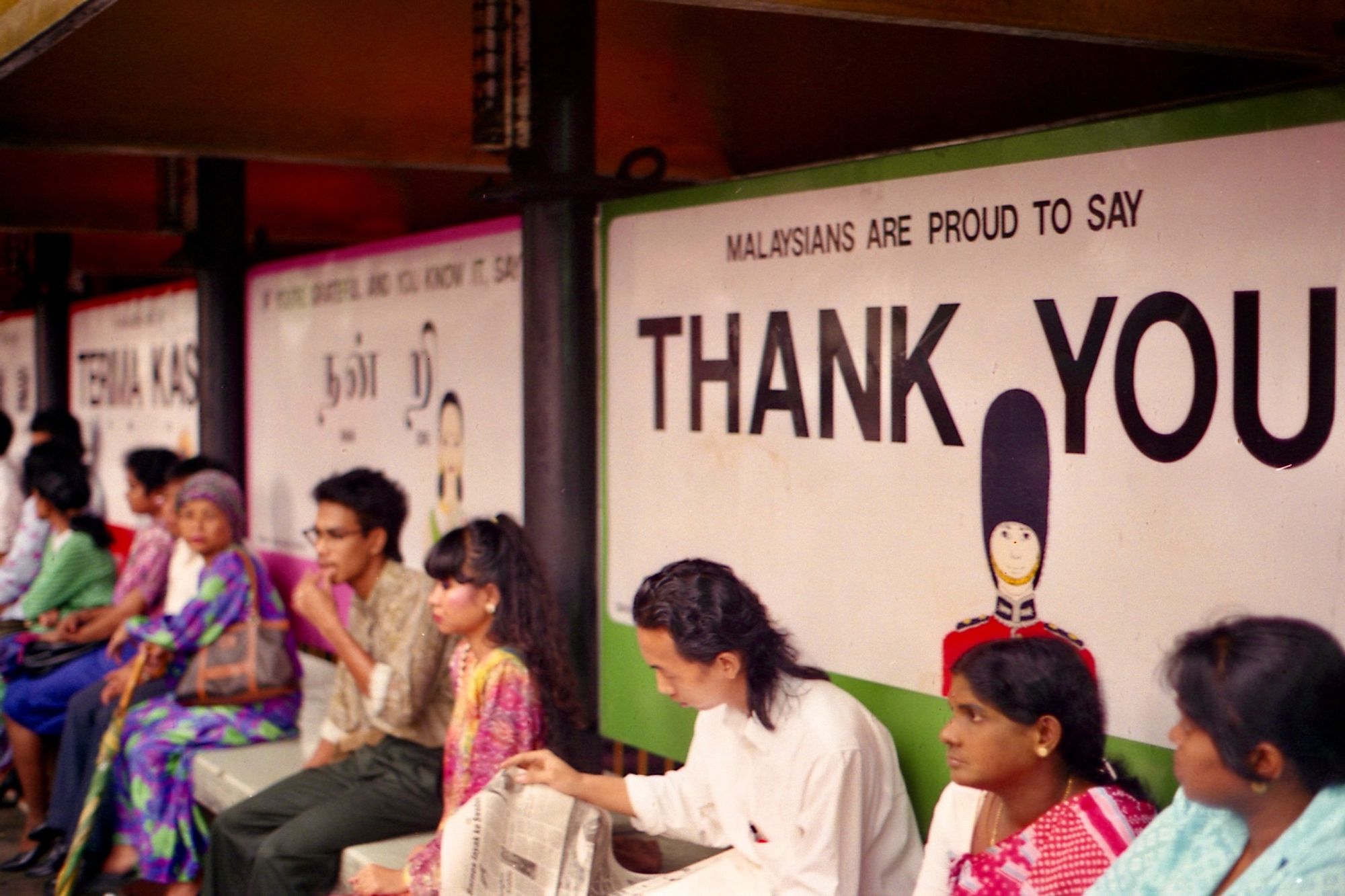 A diverse group of people sit at a bus stop. Behind them in multiple languages are signs saying ‘Malaysians are proud to say thank you’.