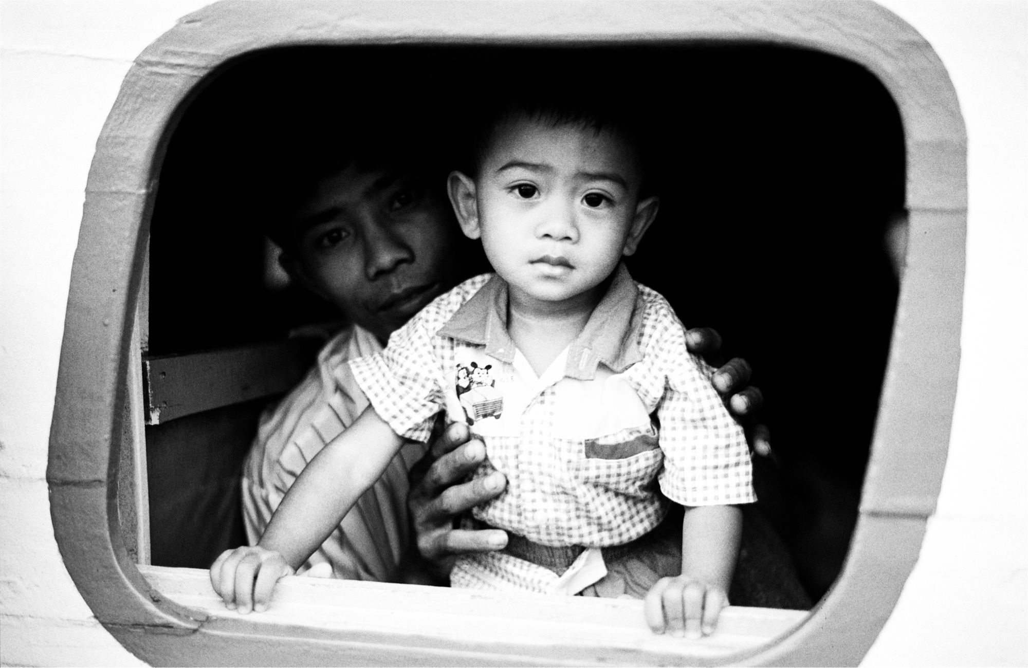 View through a ship’s porthole from the deck outside. A child is looking at the camera, behind in the shadow is a man holding the child, also looking at the camera.