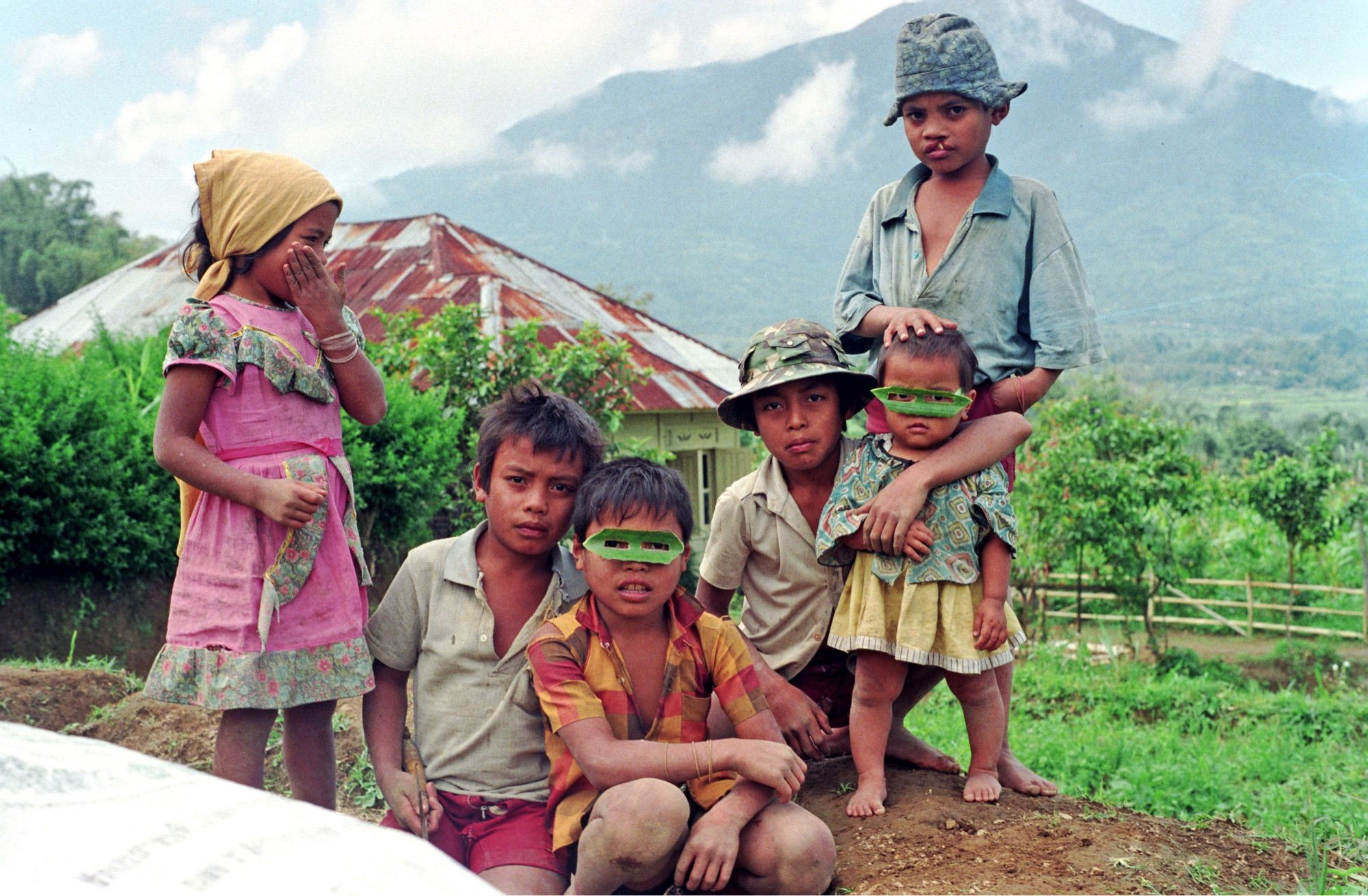 A group of six children pose squatting next to a sack of rice. Behind them is a volcano. Two of the kids wear lone ranger masks made from banana leaves. On the left a little girl is covering her laughter with her hand.