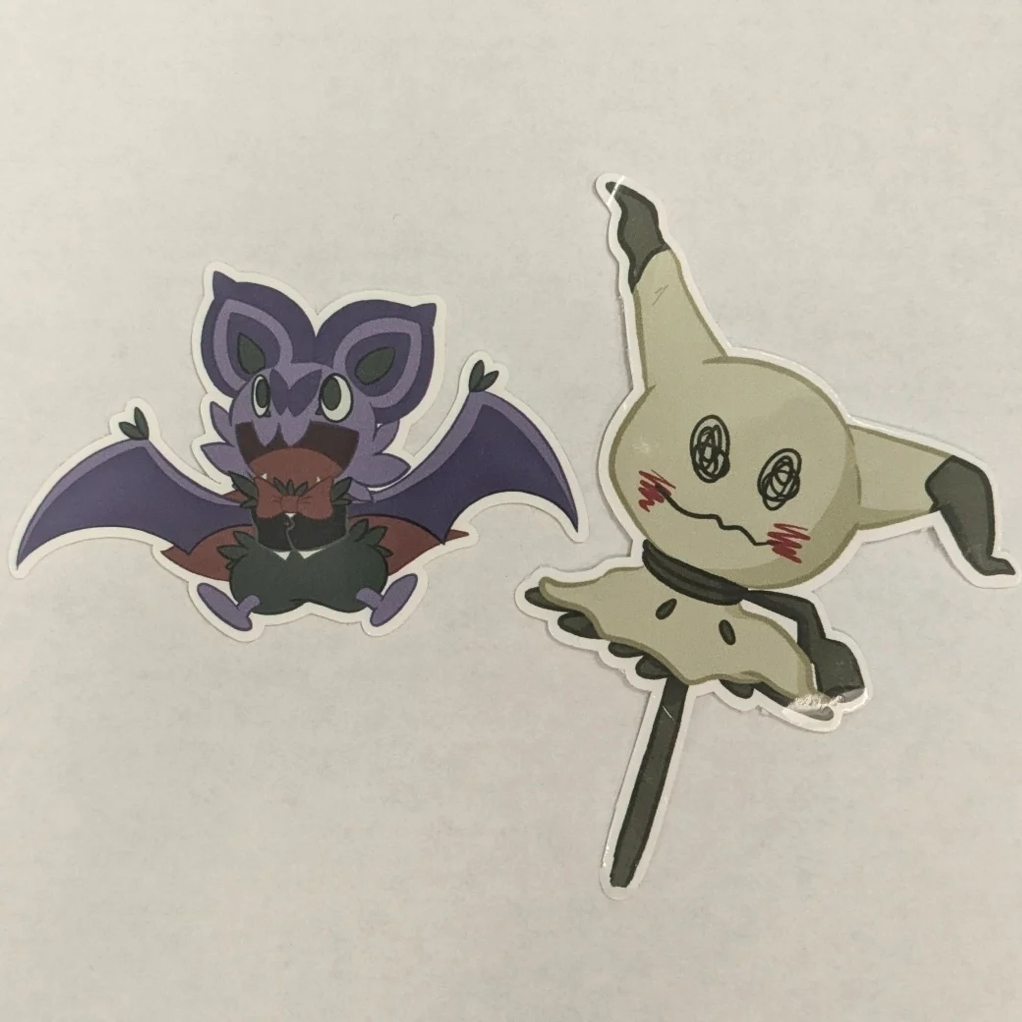 Two stickers, one of a Noibat dressed in a vampire vest, bowtie, and cape, and the other of a lollipop shaped like Mimikyu