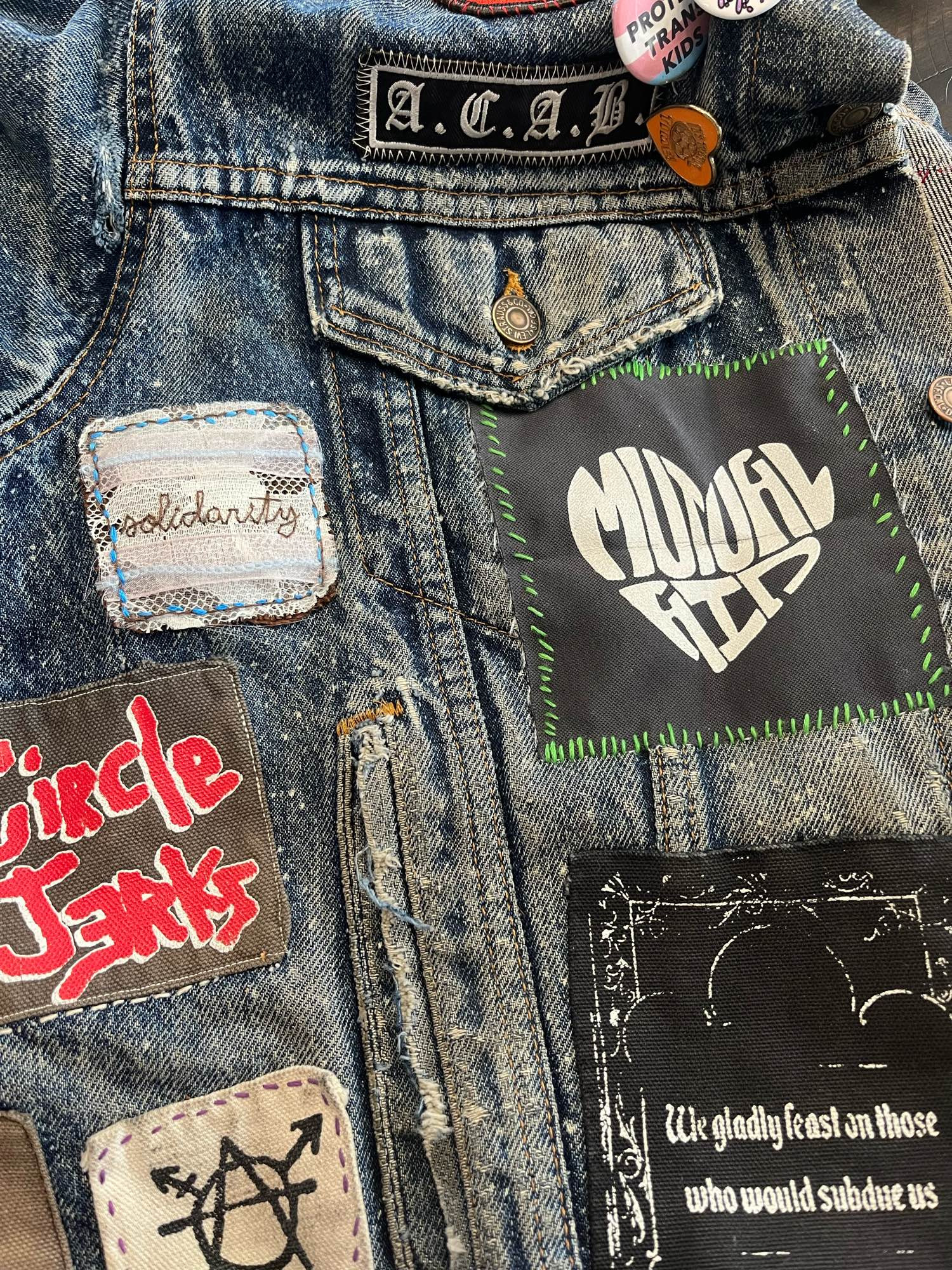 Image shows RiotDad’s jacket with several patches. One prominently displayed reads “mutual aid” in the shape of a heart in white letters in a black background. Another is handstitched pieces of fabric of miscellaneous textures that read “solidarity”, a black patch with the words “ circle jerks” in red, a white patch with the trans/gender queer anarchy symbol in black and a black patch with either letters that reads “We gladly feast upon those that would subdue us.”