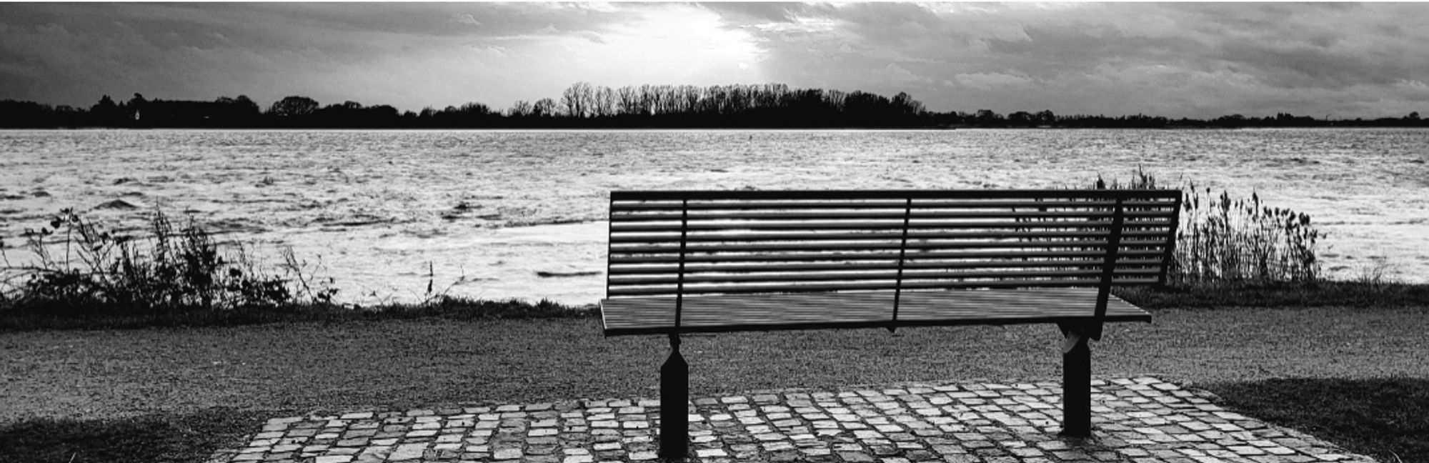 Eine Bank steht auf gepflastertem Boden, dahinter ein befestigten Sandweg, dahinter ein Flusss, am Horizont das gegenüberliegende Ufer, der Himmel kündigt bereits Regen an