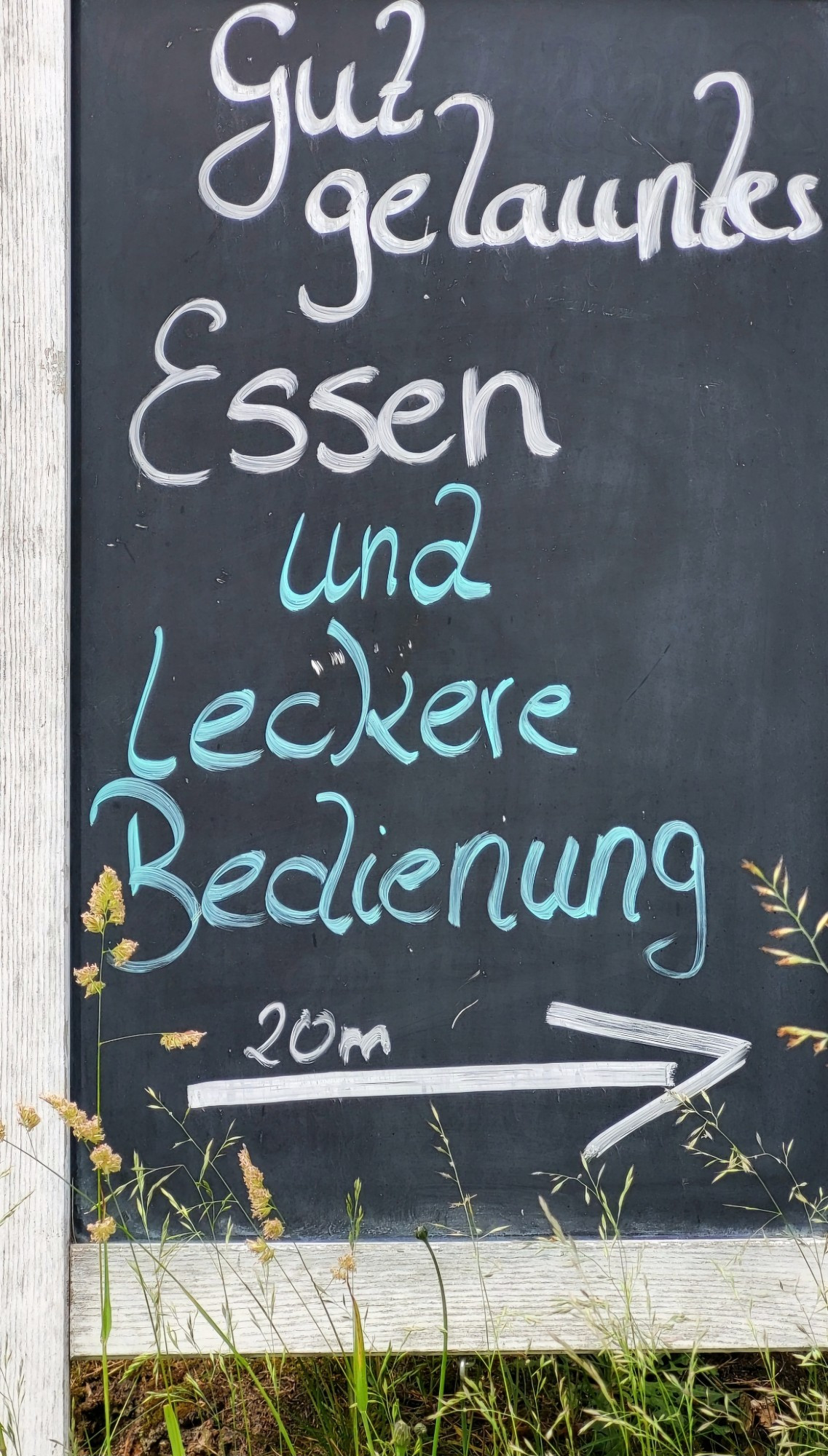 Hochkant über das volle Format eine in Holz eingefasste Standtafel mit handschriftlichem Text in weiß/türkis: gut gelauntes Essen und leckere Bedienung. Darunter ein Pfeil nach rechts und der Hinweis 20 m. Unterhalb der Standtafel wachsen Gräser.