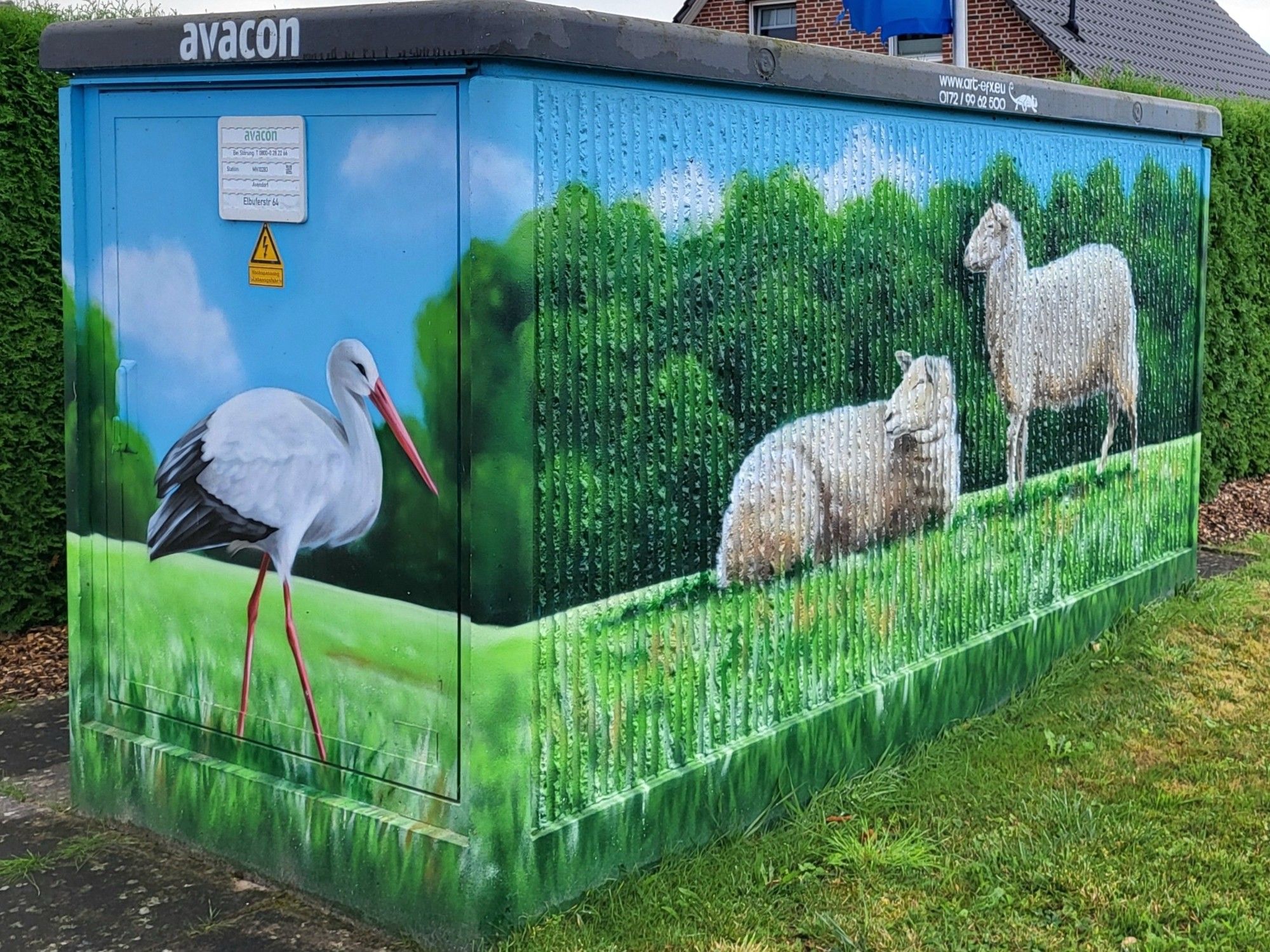 Ein verschönert Trafohäuschen. Links auf der schmalen Seite ein Storch auf grüner Wiese, schreitet von links nach rechts. Die grüne Wiese setzt sich auf der breiten Seite fort. Hier befinden sich zwei Schafe, liegend und stehend vor Büschen, die nach links Richtung Storch schauen. Hellblauer Himmel mit Wölkchen.