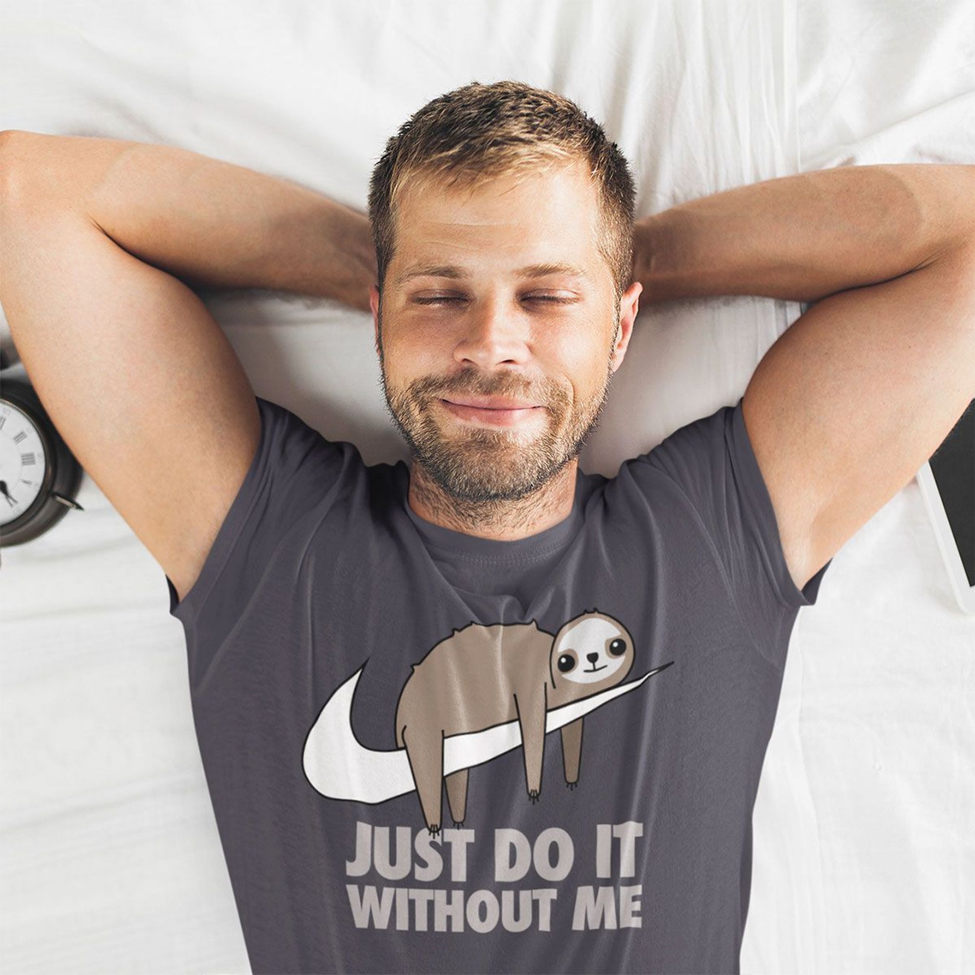 A man in a grey t-shirt is lying on his bed. On the front of the T-shirt is a picture of a sloth lying down over a swoosh and the words "Just do it without me" underneath.