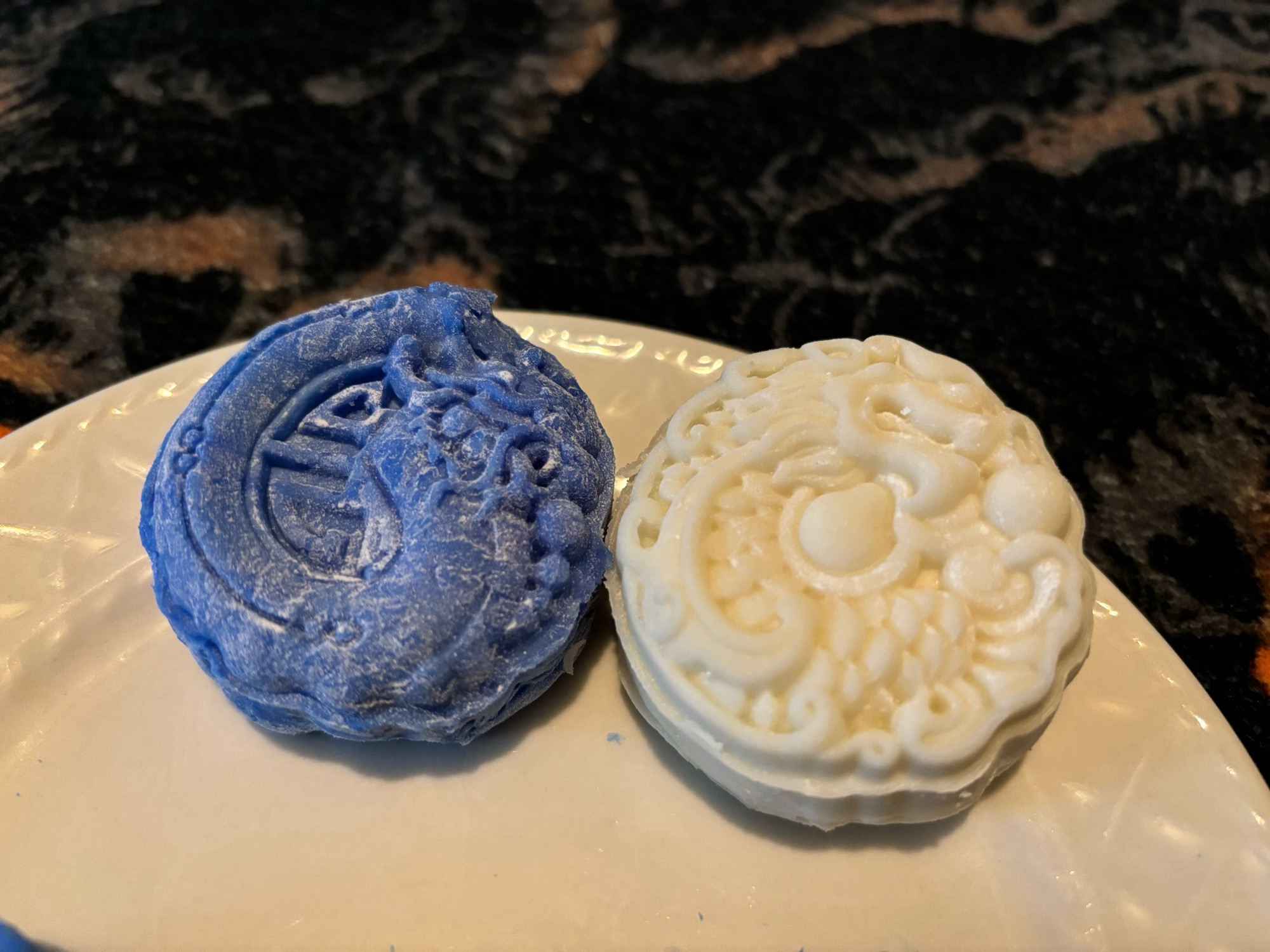 Two snow skin moon cakes, one blue and one white/uncolored. Both mooncakes have similar eastern dragon designs with some alterations: the blue one has a lucky symbol in the background and the white has dragon pearls.