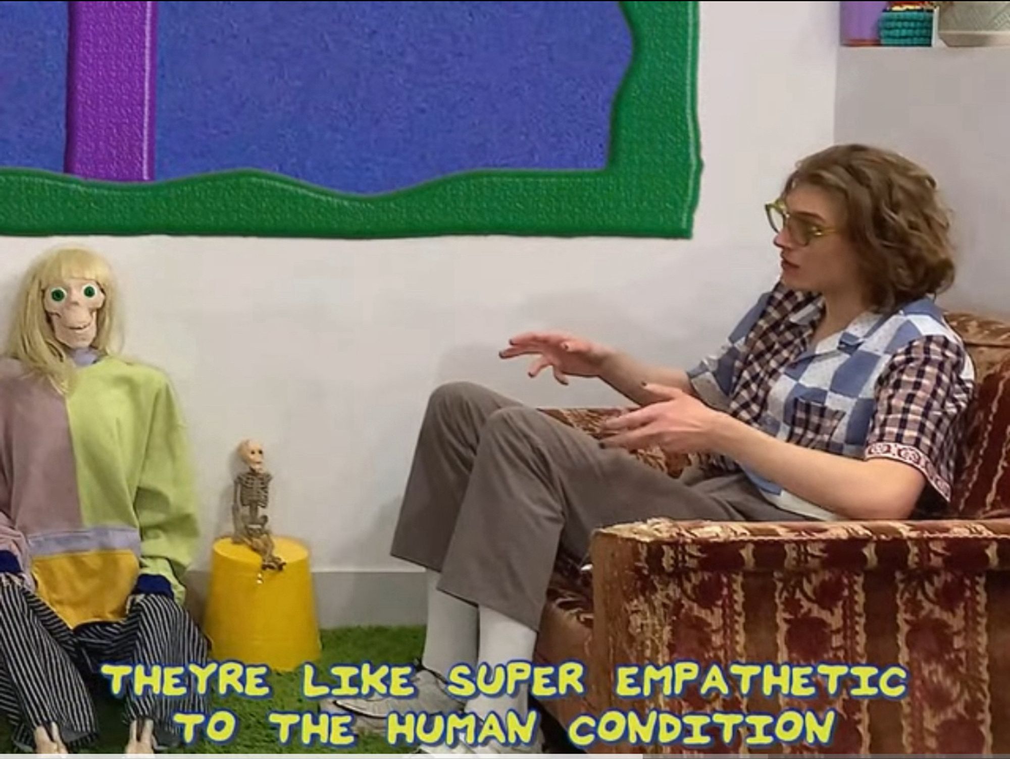 Still from drive45 featuring a nonbinary individual sitting in a chair in a cartoony looking room that also has two skeletons in it, one is wearing clothes. The subtitles read “They’re like super empathetic to the human condition”