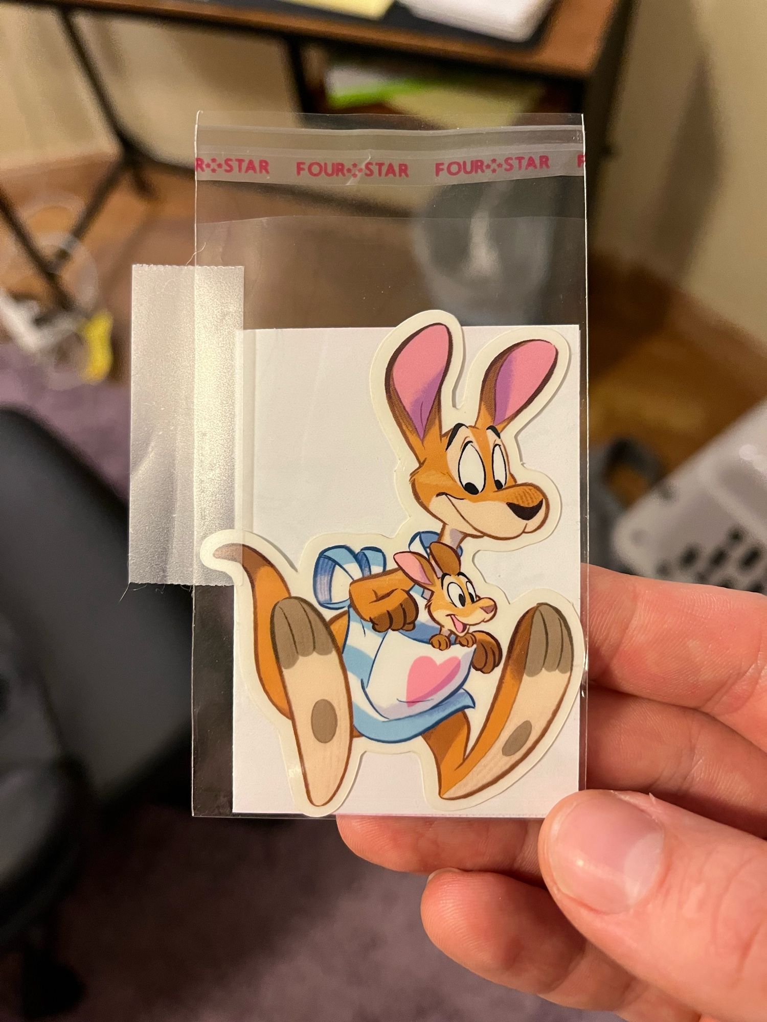 Sticker of a toony kangaroo wearing a blue and white apron with the big pouch where the pouch goes, there’s a Joey in there who’s excited to be along for the ride