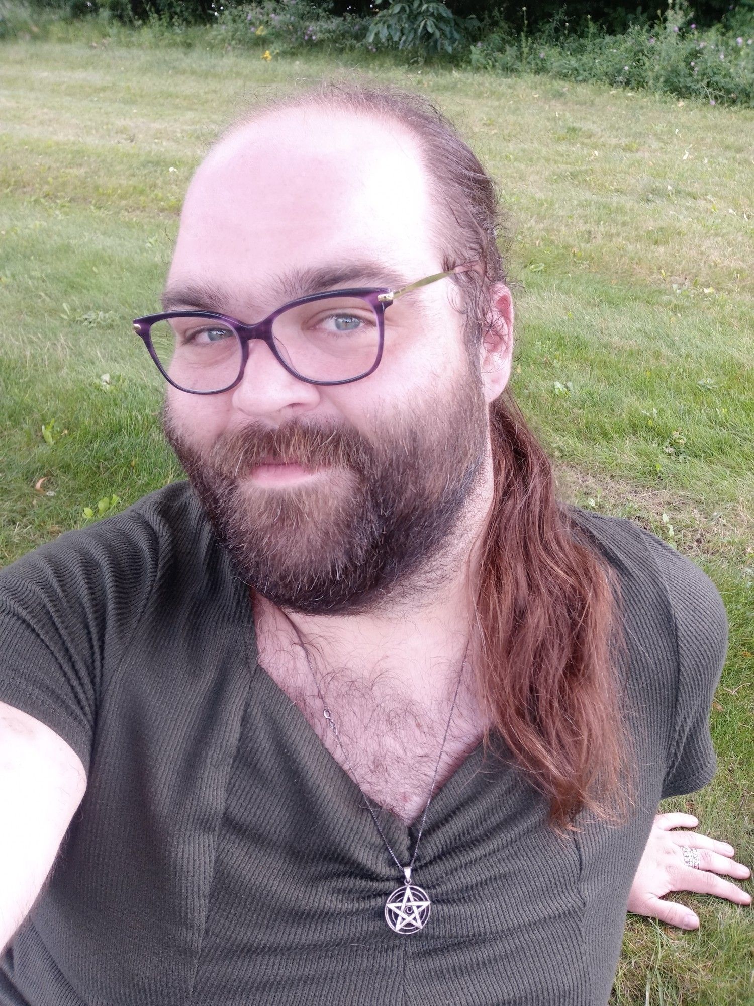 Me! A transgender woman with a thick, short cut beard and a big forehead. She's wearing glasses and a pentagram.