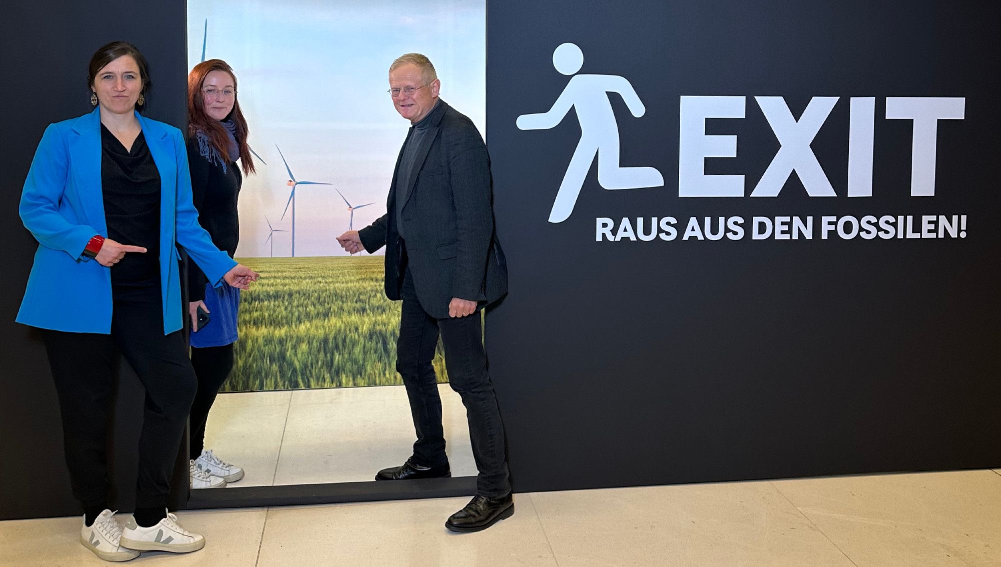 Auf dem Bild erkennt man unsere Abgeordneten Katharina Beck, Kathrin Henneberger und Armin Grau. Sie stehen in einem stilisierten Türrahmen einer schwarzen Plakatwand. Auf der Wand steht in weißer Schrift "EXIT - Raus aus den Fossilen". Durch den Türausschnitt erkennt man eine Landschaft mit Windrädern.