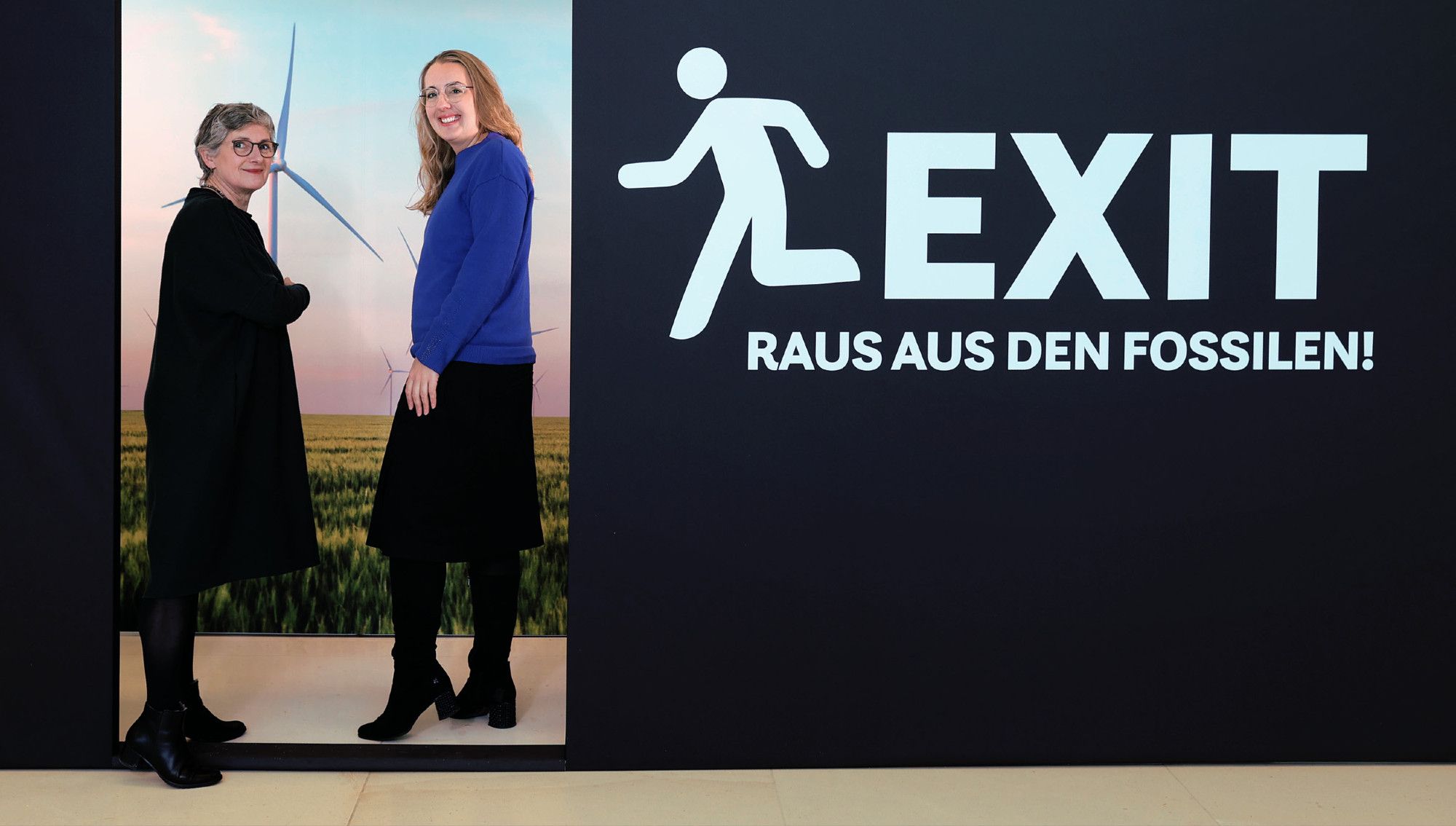 Auf dem Bild erkennt man unsere Fraktionsvorsitzenden Britta Haßelmann und Katharina Dröge. Sie stehen in einem stilisierten Türrahmen einer schwarzen Plakatwand. Auf der Wand steht in weißer Schrift "EXIT - Raus aus den Fossilen". Durch den Türausschnitt erkennt man eine Landschaft mit Windrädern.