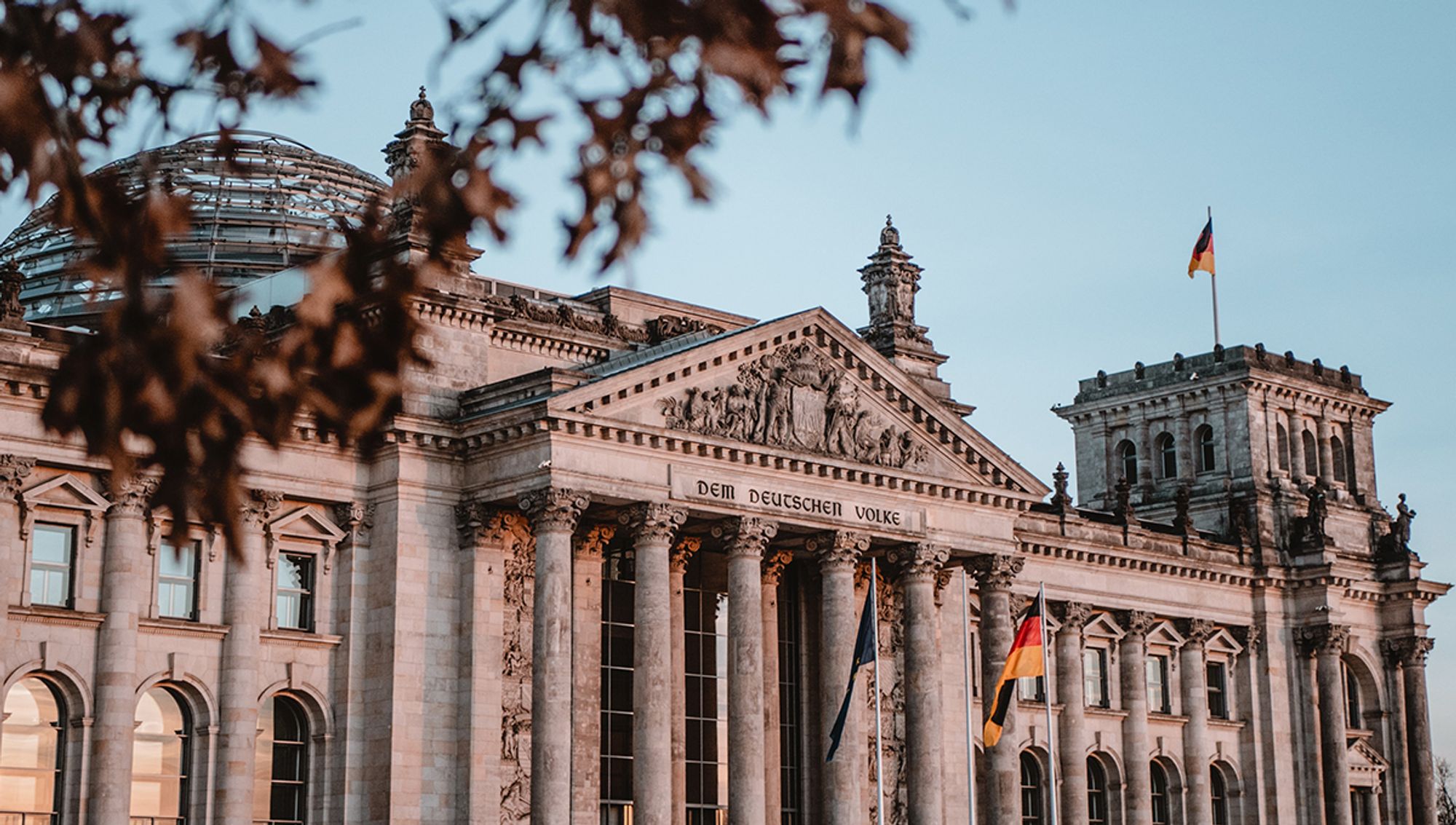 Auf dem Foto ist der Reichstag zu sehen. Im Vordergrund sind verschwommen herbstliche Blätter zu erkennen.