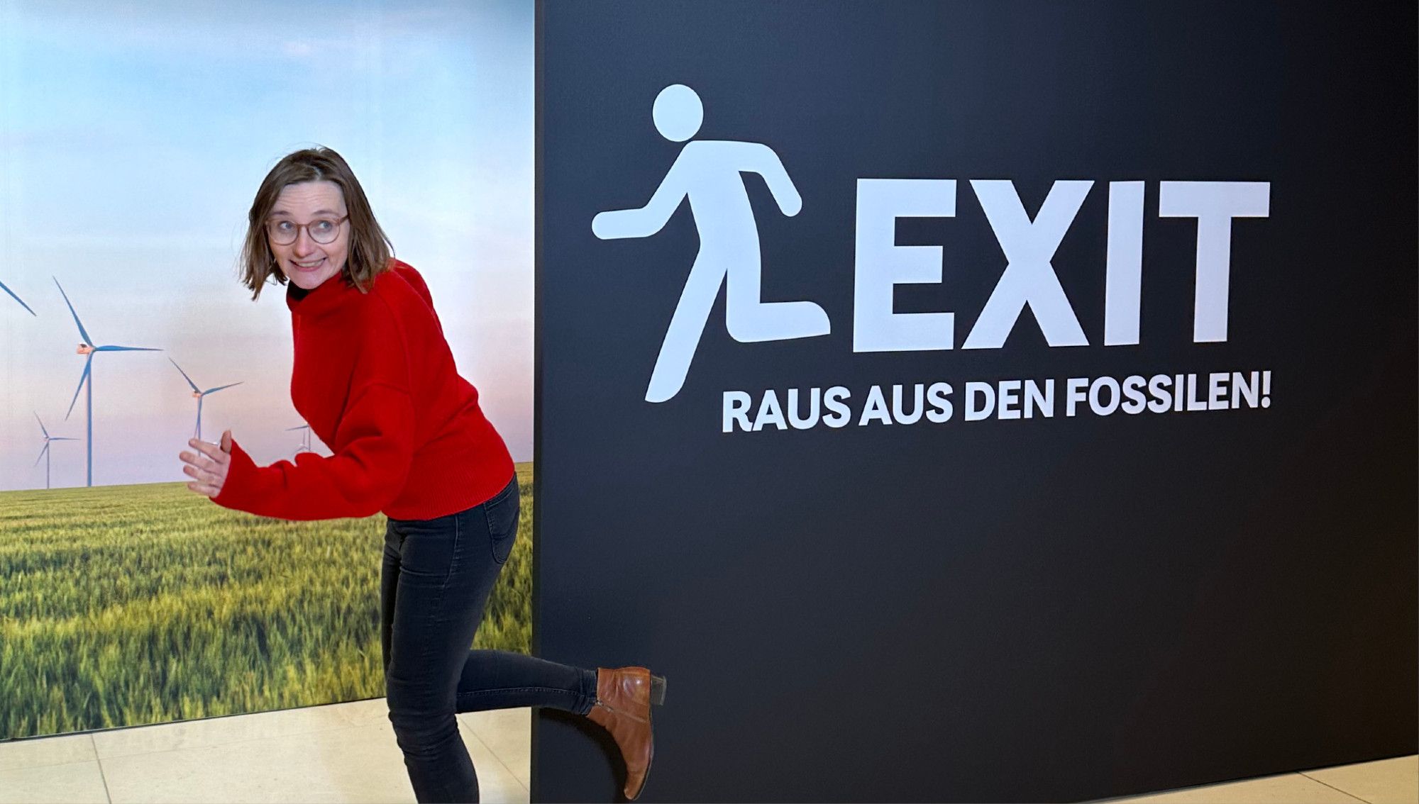 Auf dem Bild erkennt man unsere Abgeordnete Lisa Badum. Sie geht durch einen stilisierten Türrahmen einer schwarzen Plakatwand. Auf der Wand steht in weißer Schrift "EXIT - Raus aus den Fossilen". Durch den Türausschnitt erkennt man eine Landschaft mit Windrädern.