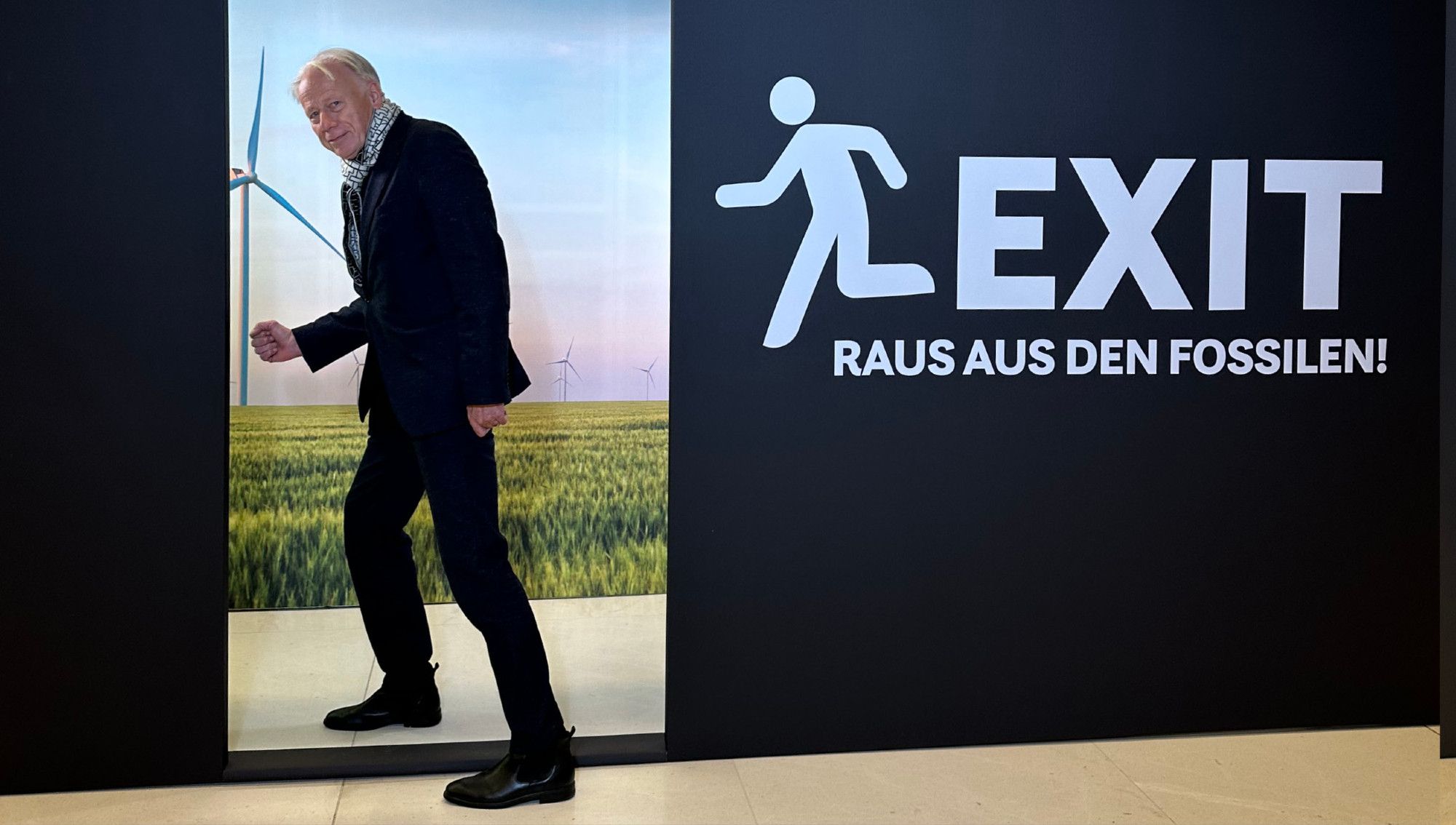 Auf dem Bild erkennt man unseren Abgeordneten Jürgen Trittin. Er geht durch einen stilisierten Türrahmen einer schwarzen Plakatwand. Auf der Wand steht in weißer Schrift "EXIT - Raus aus den Fossilen". Durch den Türausschnitt erkennt man eine Landschaft mit Windrädern.