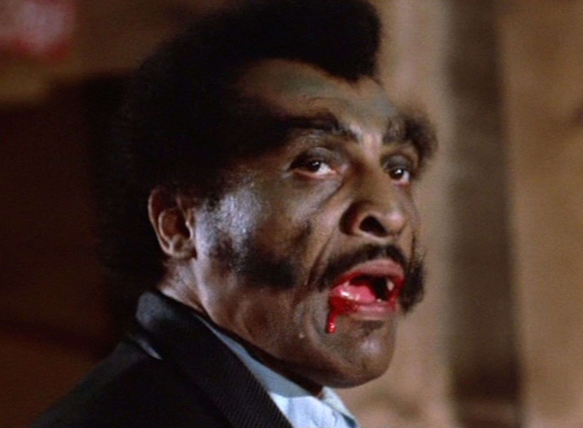 William Marshall as Blacula, licking his lips dripping with blood.  he sports a wild mustache/horizontal cheek sideburns for facial hair and bushy eyebrows.  his skin has a greenish tint.  Marshall was also the King of Cartoons on Pee Wee's Playhouse