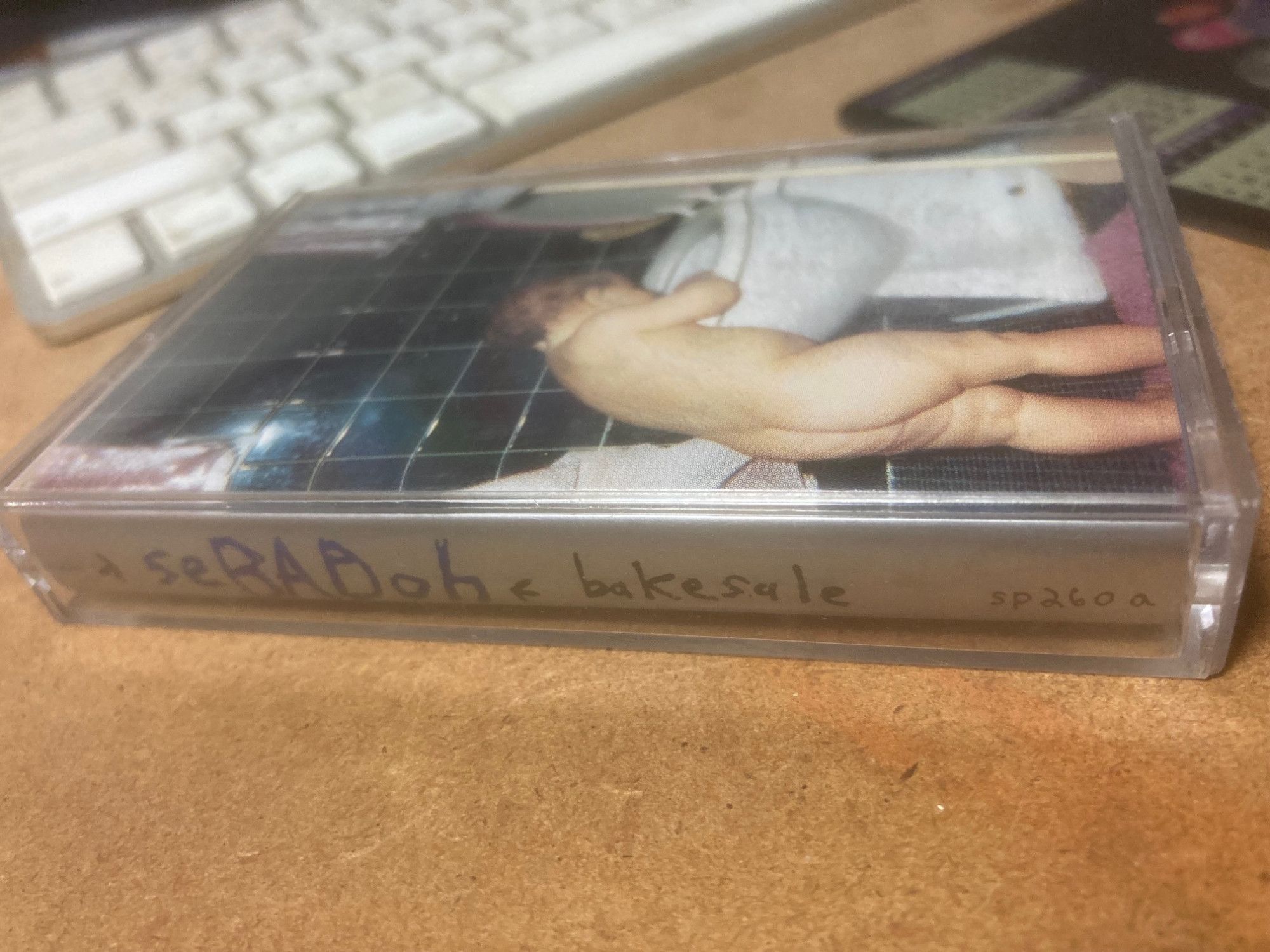 a clear cassette tape case with inside printed card with Sebadoh bakesale in blue type on solid silver background on spine and a picture of a naked butt baby standing with hands on rim of a toilet looking in which was the cover of the album.  it sits on my desk and my keyboard is in short distance background