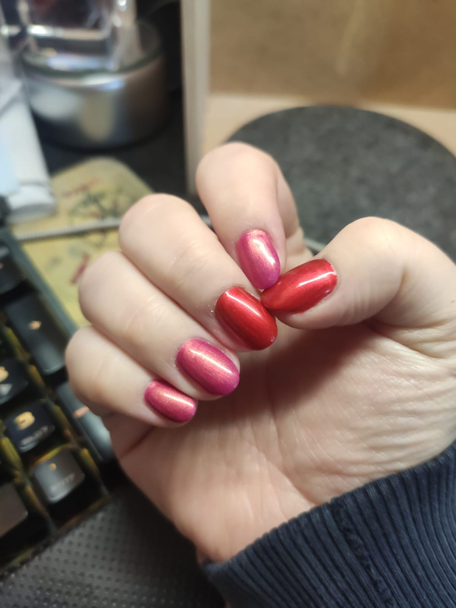 Das Bild zeigt eine Nahaufnahme einer Hand mit lackierten Fingernägeln. Die Nägel sind in zwei verschiedenen Farben lackiert: Der Daumen, der Zeigefinger und der kleine Finger haben eine metallisch-rote Farbe, während der Mittel- und der Ringfinger in einem metallisch-pinken Ton lackiert sind.