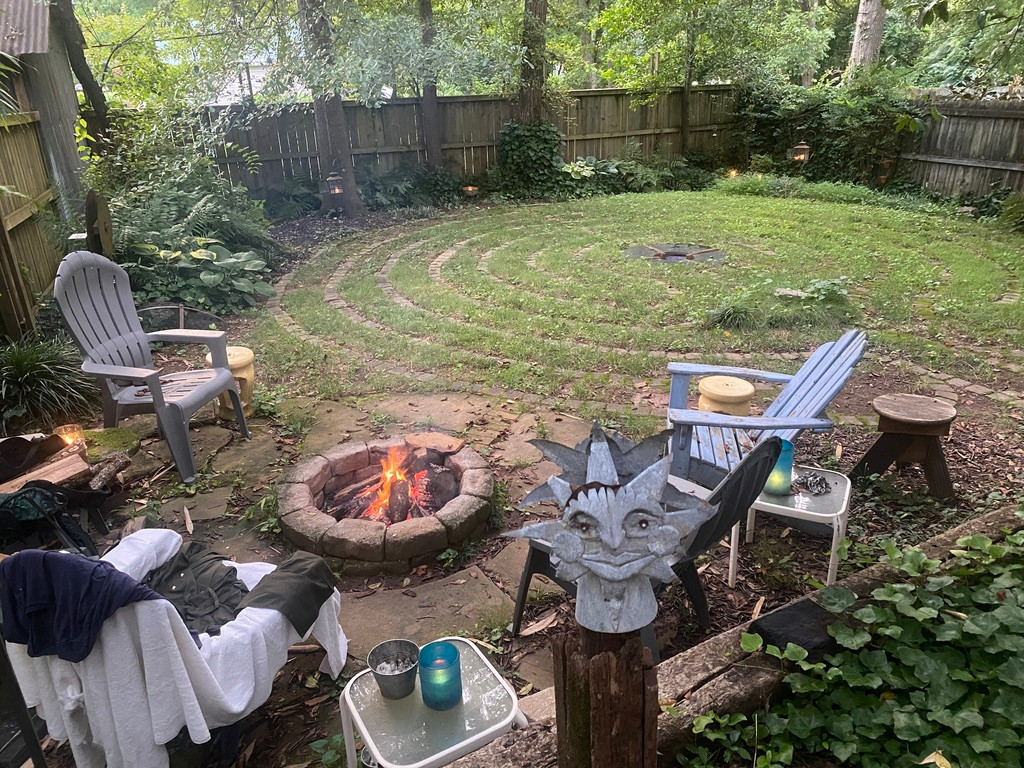 a 7-circuit labyrinth in a back yard. In the lower left foreground is a fire pit (with a fire lit in it) surrounded by Adirondack chairs. The labyrinth, a grassy path outlined with paving stones, is in the upper left of the shot. It is an altogether serene place.