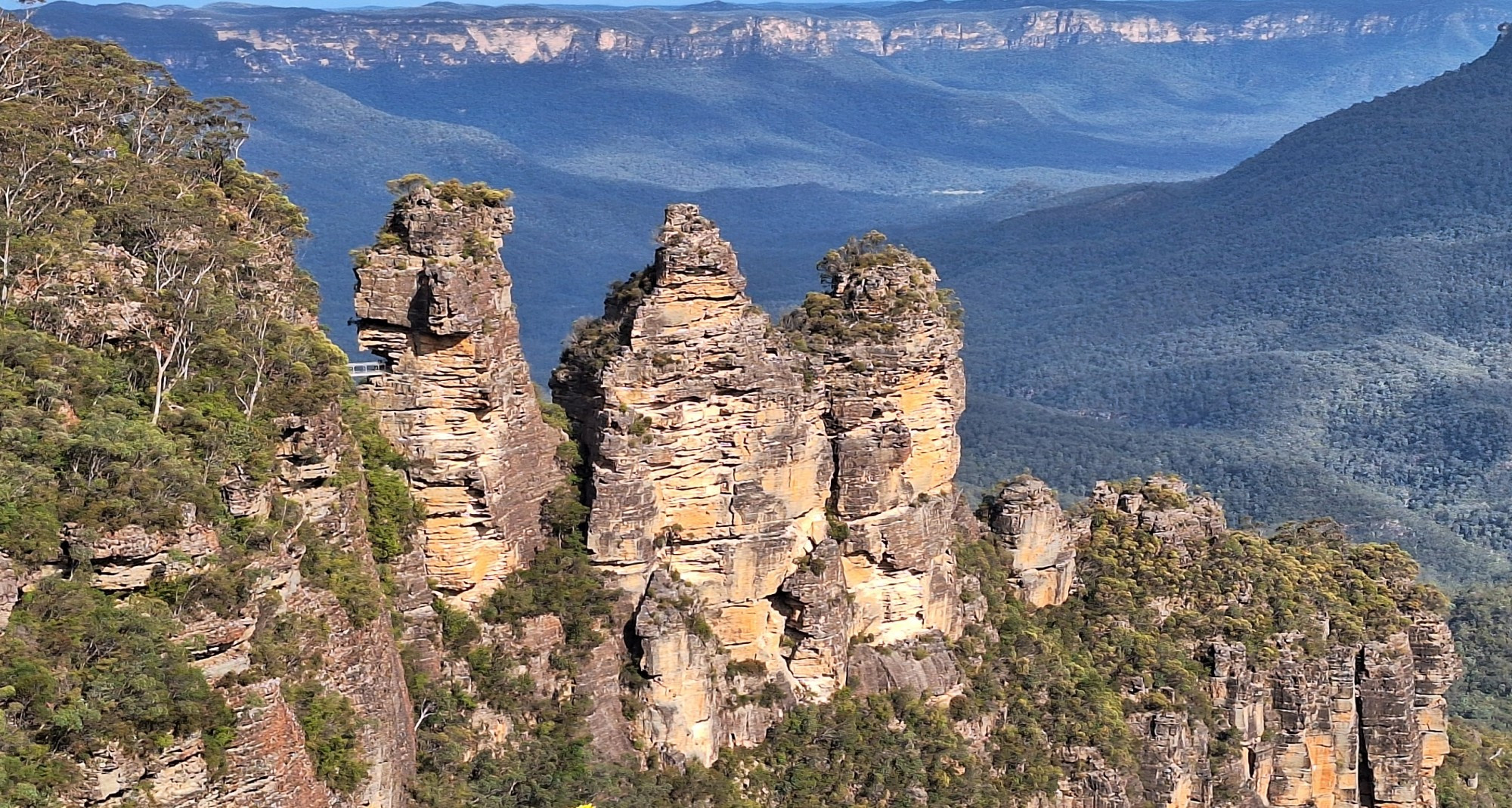 "Three Sisters", Katoomba, Blue Mountains, New South Wales, Australia:
large sunlit rocks in the foreground, rocky cliffs in distant background, wide Canyon filled with green trees between