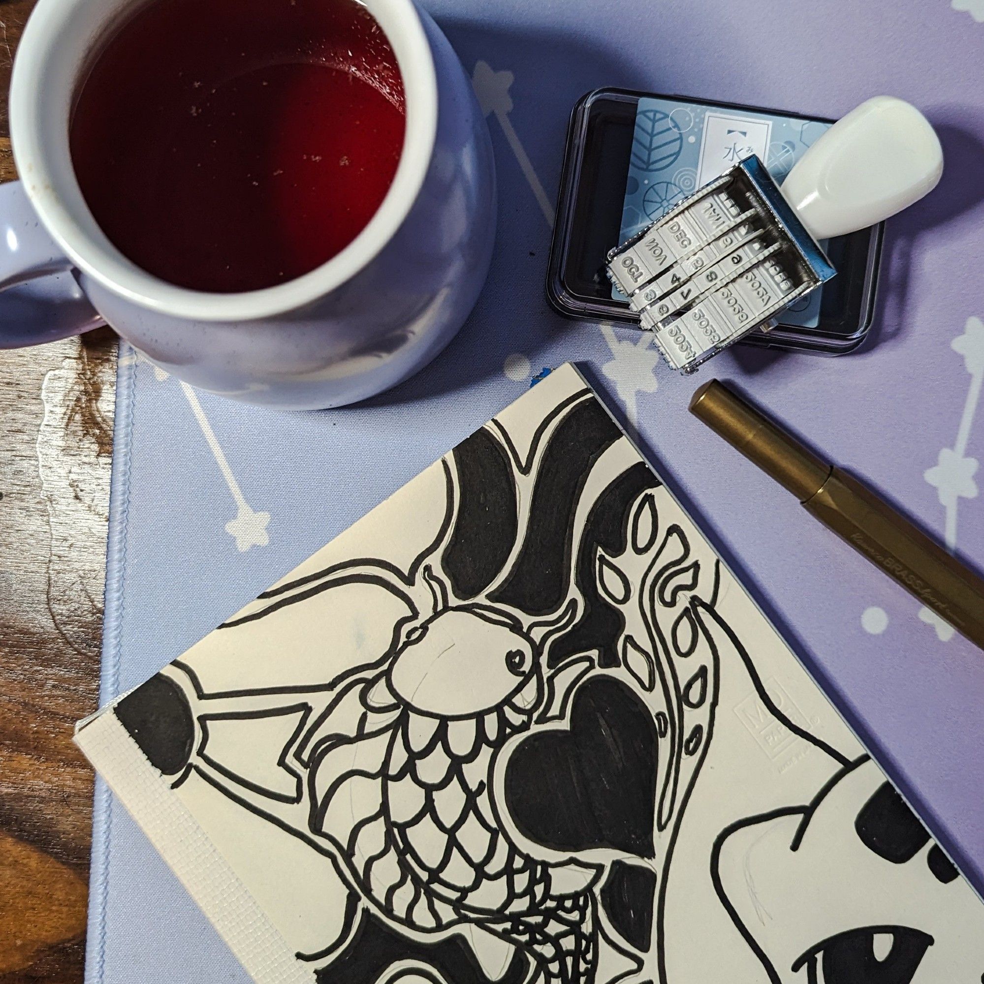 Overhead photo of a purple coffee mug filled with tea, a date stamper and blue ink pad, a brass fountain pen, and a journal.