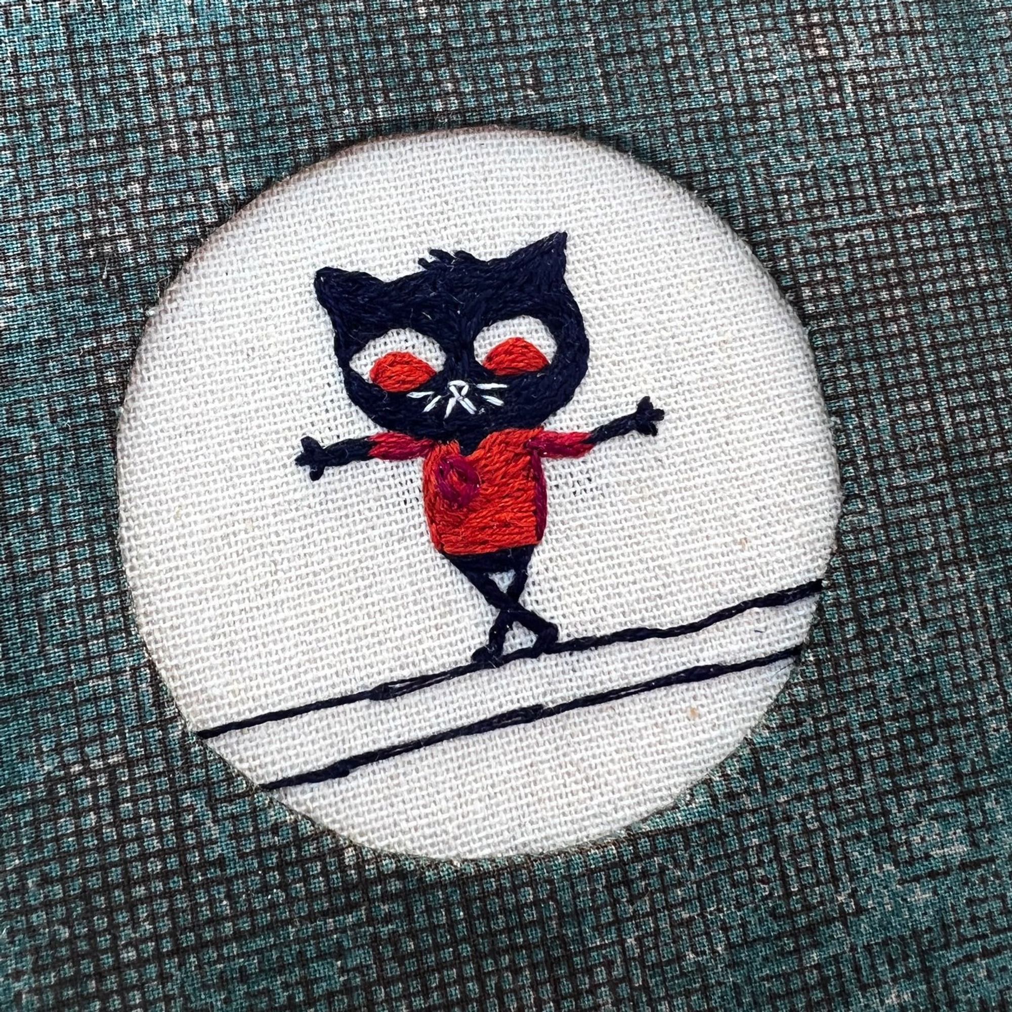 close up image of the Mae (character from Night in the Woods game) embroidery embedded into the cover. She is a black cat walking on a power line with her arms outstretched, wearing her signature orange-y reddish shirt. She is watching her steps.