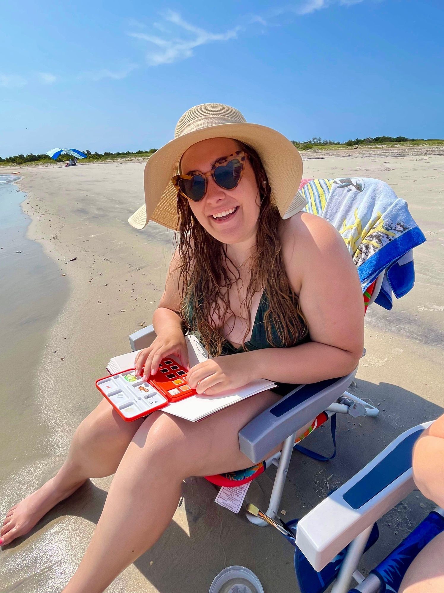 a photo of liz smiling, sitting on a beach chair at the water’s edge at the shore of shackleford banks. she is wearing heart shaped brown sunglasses, a floppy beige hat, and a deep v cut dark green swimsuit. her long brown hair is damp and she is in the process of setting up her outdoor watercolor paint set in her lap.