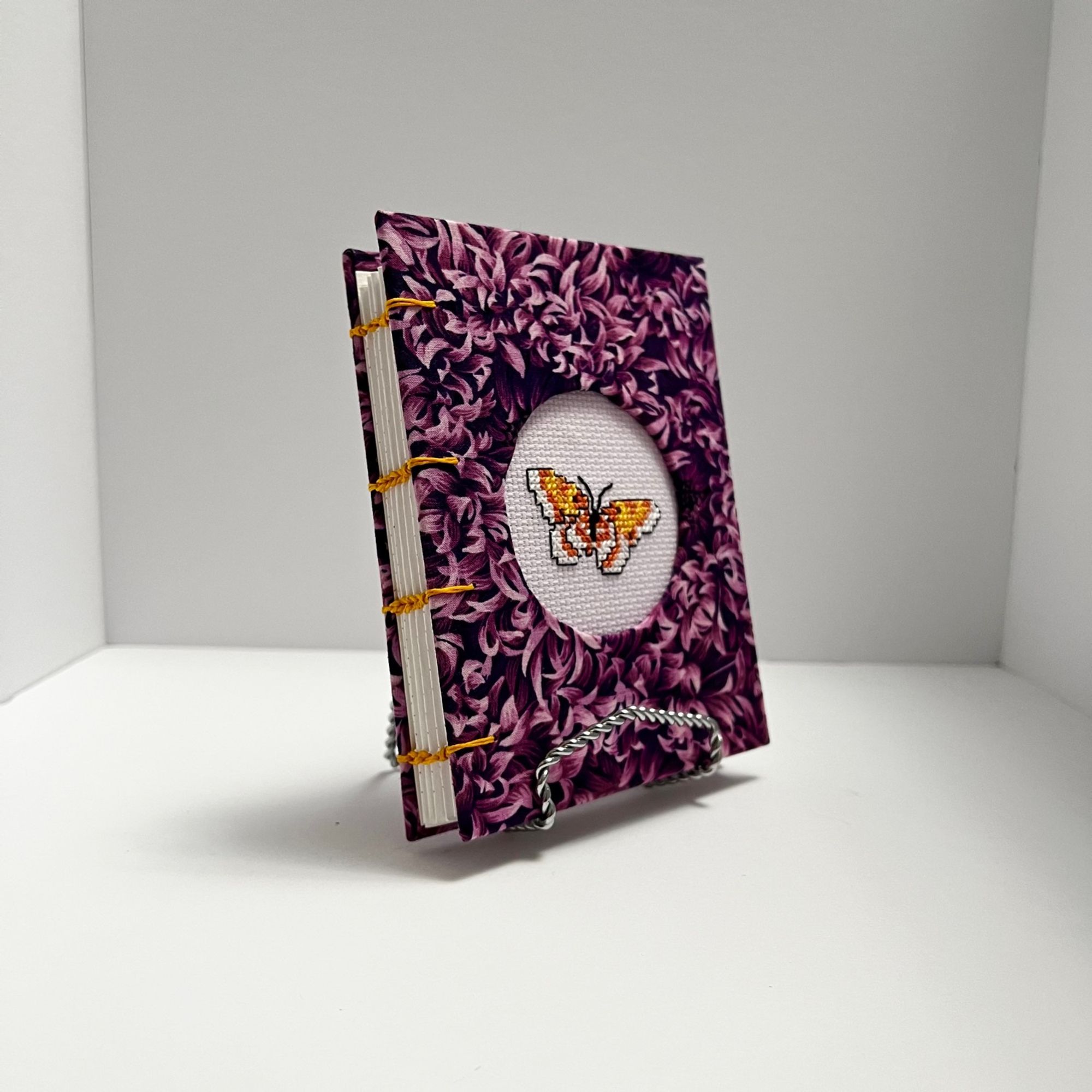 a 3/4 angle view of a hand bound coptic stitch journal with purple petal pattern covers. there is an orange, white, and black butterfly cross stitch features in the cover.