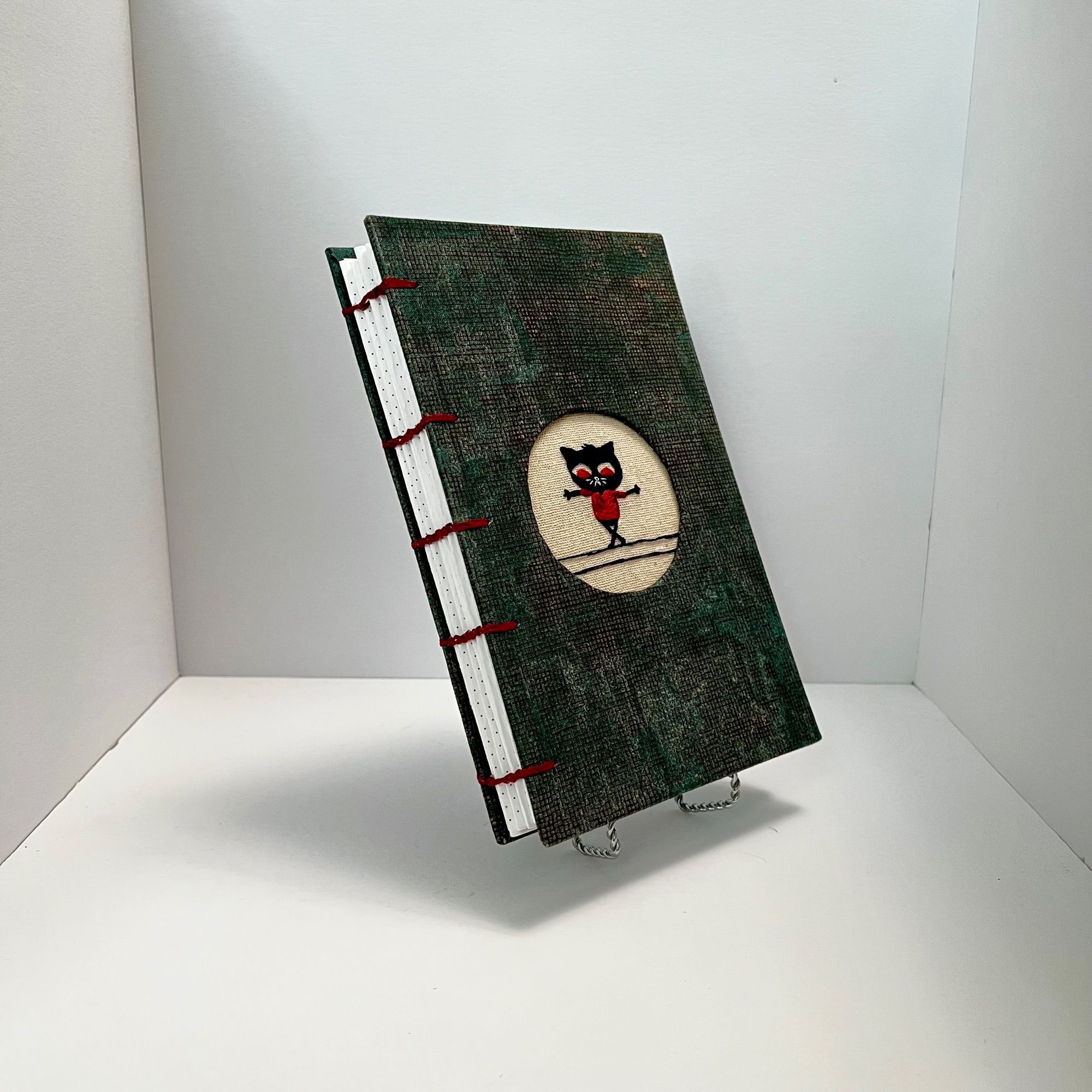 3/4 angle view of a hand bound coptic stitch journal with dark green covers and dark red stitching on the spine. White background, there is a Mae (character from Night in the Woods game) embroidery embedded into the cover.