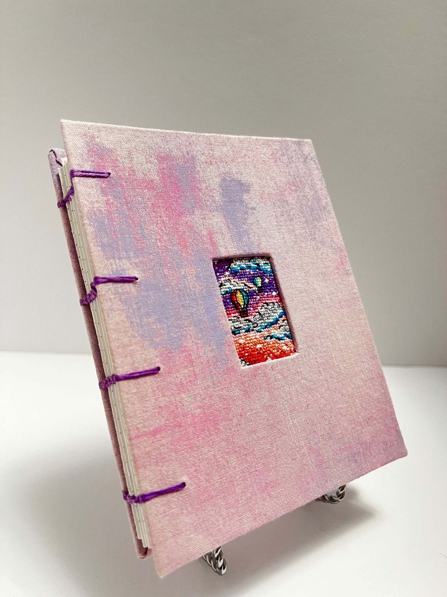 hand made coptic stitch notebook with light pink and purple covers, with a cross stitch of hot air balloons at sunset embedded into the cover