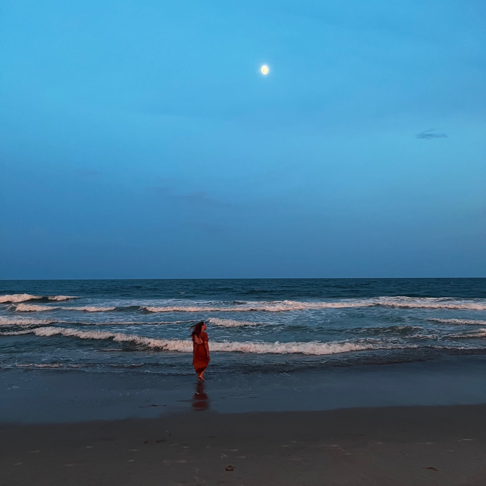 a photo of liz in the distance, just after sunset, walking along the ocean waves under a moon. she is wearing a two piece mustard orange wrap skirt and top set.