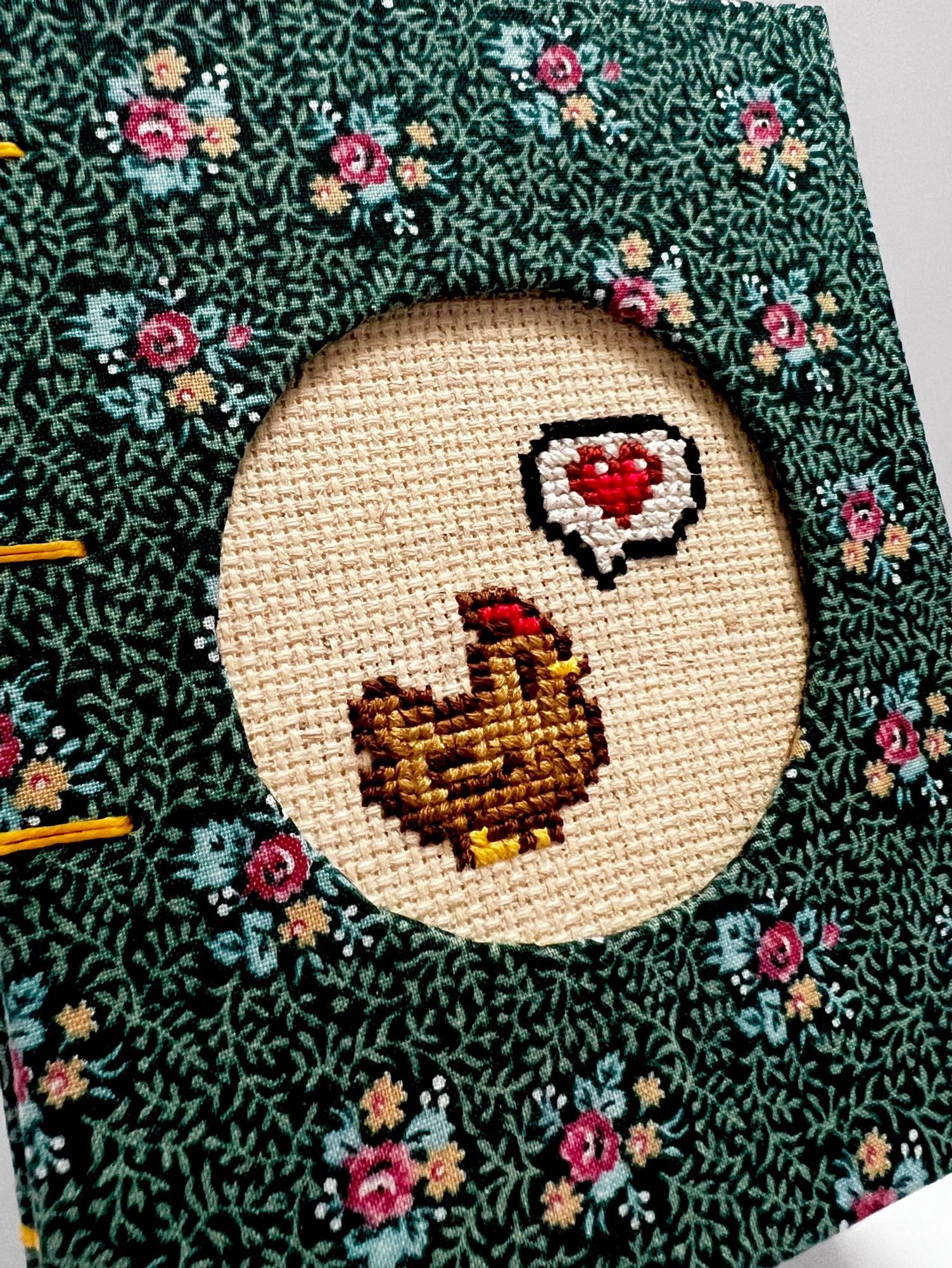 close up image of a cross stitch of a stardew valley chicken embedded into the cover of a hand made journal with a dark green floral cover