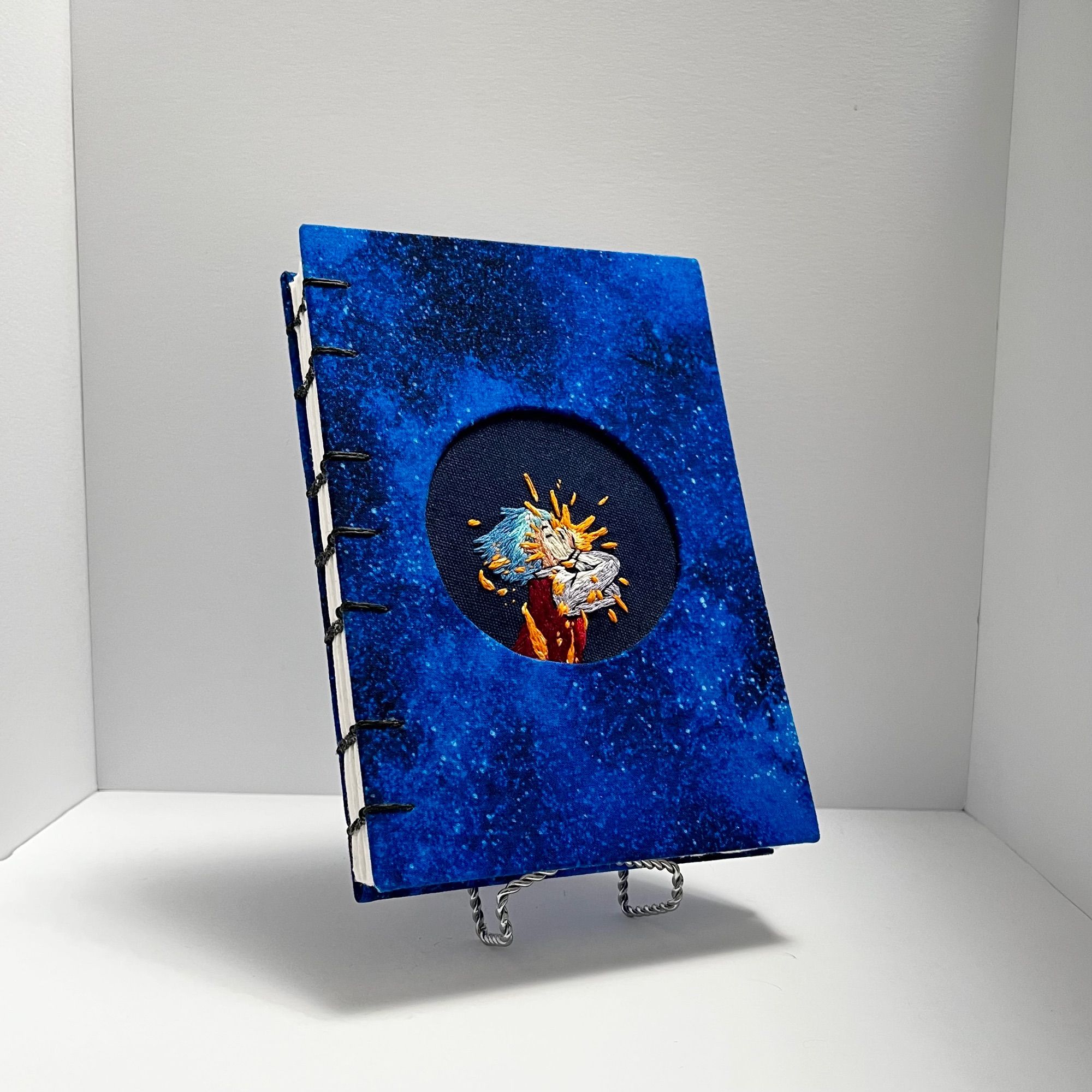 hand made coptic stitch notebook with dark blue galaxy covers and an embroidery inspired by Howl’s moving castle embedded into the cover. The embroidery design is of Howl eating the star.