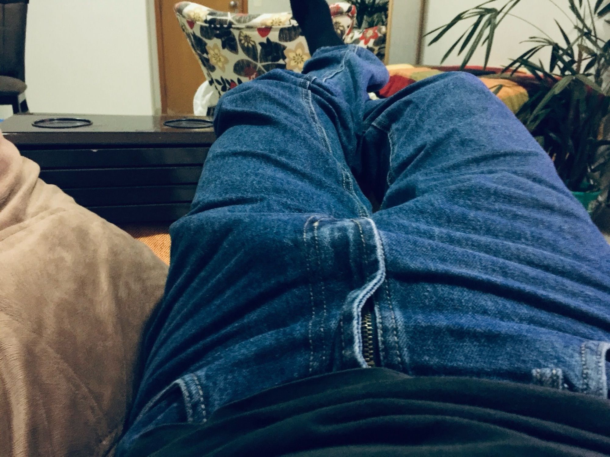 A man wearing blue jeans is lying on a couch. He also wears socks and it's noticeable a bulge on his crotch under his jeans, suggesting he's having an erection.