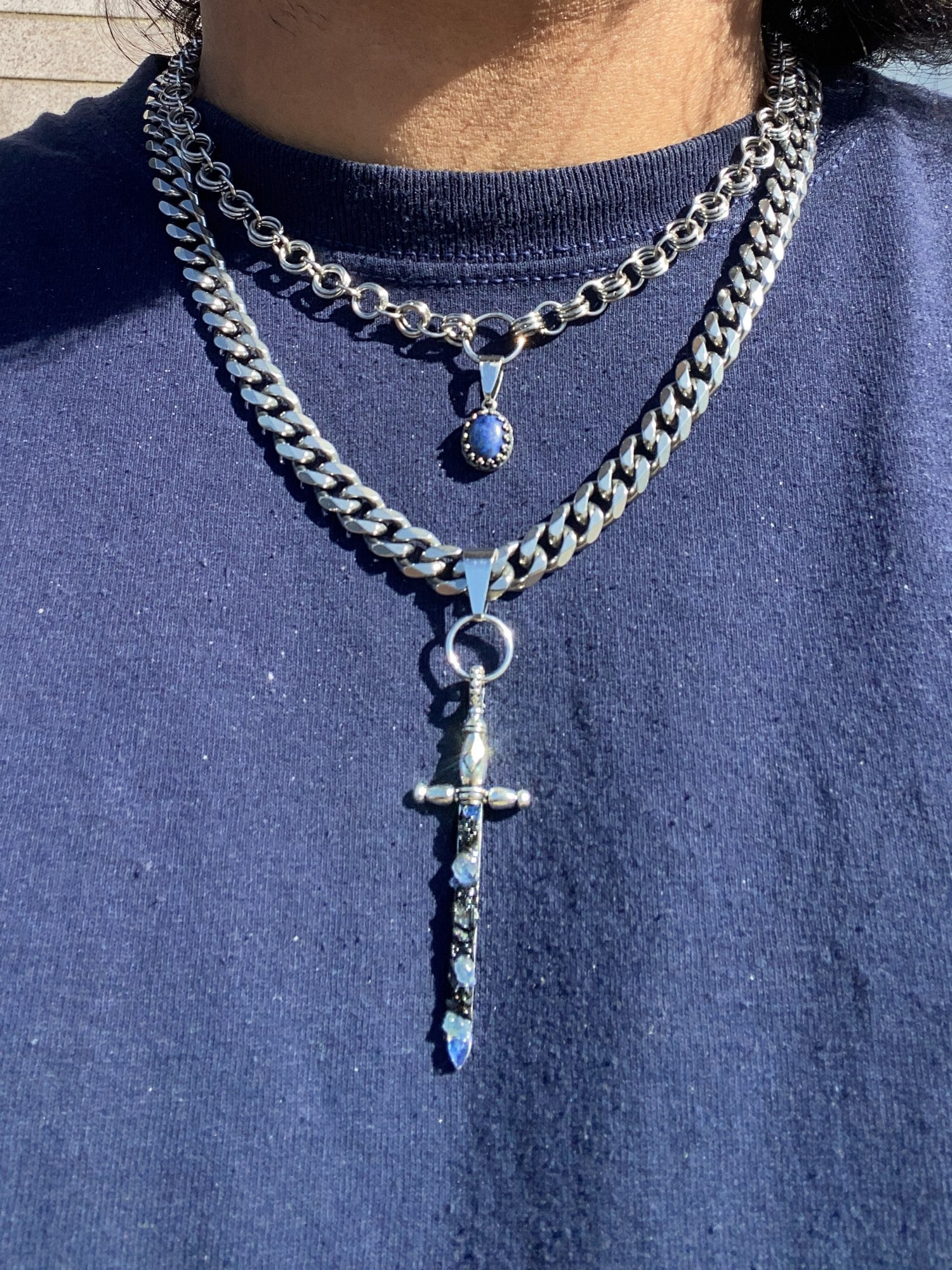 Close up of someone wearing two necklaces, the smaller one made of interlinking hoops with Oval Lapis Lazuli Pendant. Second Longer Cuban Link Chain Necklace has a Sword Pendant with Sapphire and Black Tourmaline.