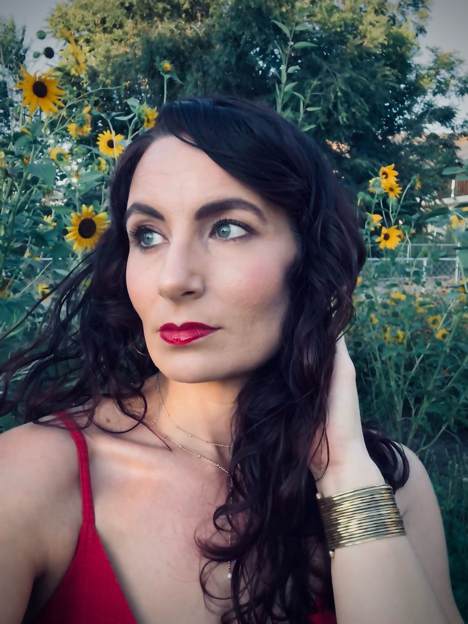 A selfie of the author. They have dark curly hair, green eyes, looking off to the distance and red lips. They’re wearing a red dress and - gold bracelet. There are sunflowers in the background.