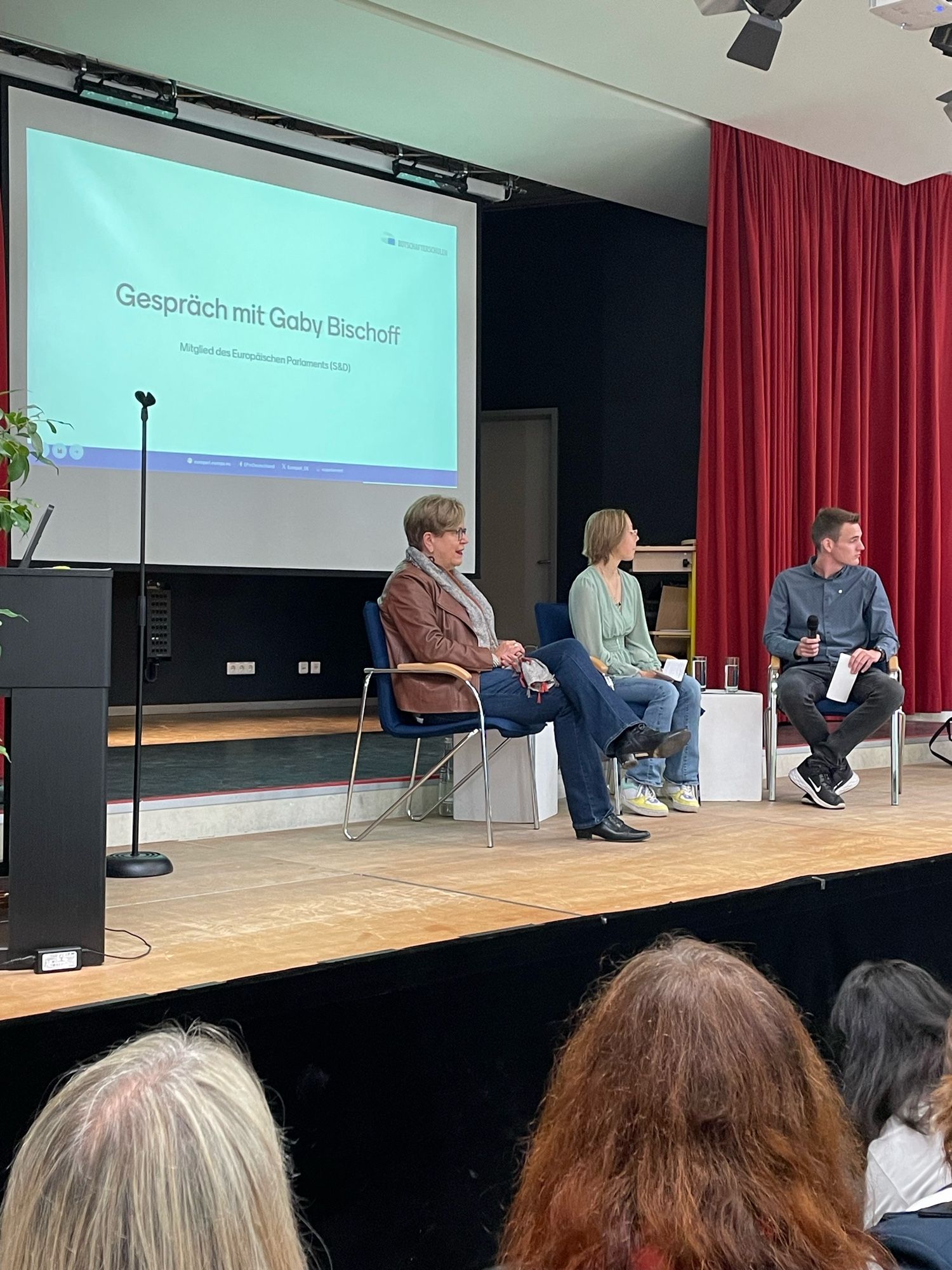 Das Podium auf der Bühne ist eröffnet. Zwei Schüler:innen sitzen gemeinsam mit Gaby Bischoff auf der Bühne.