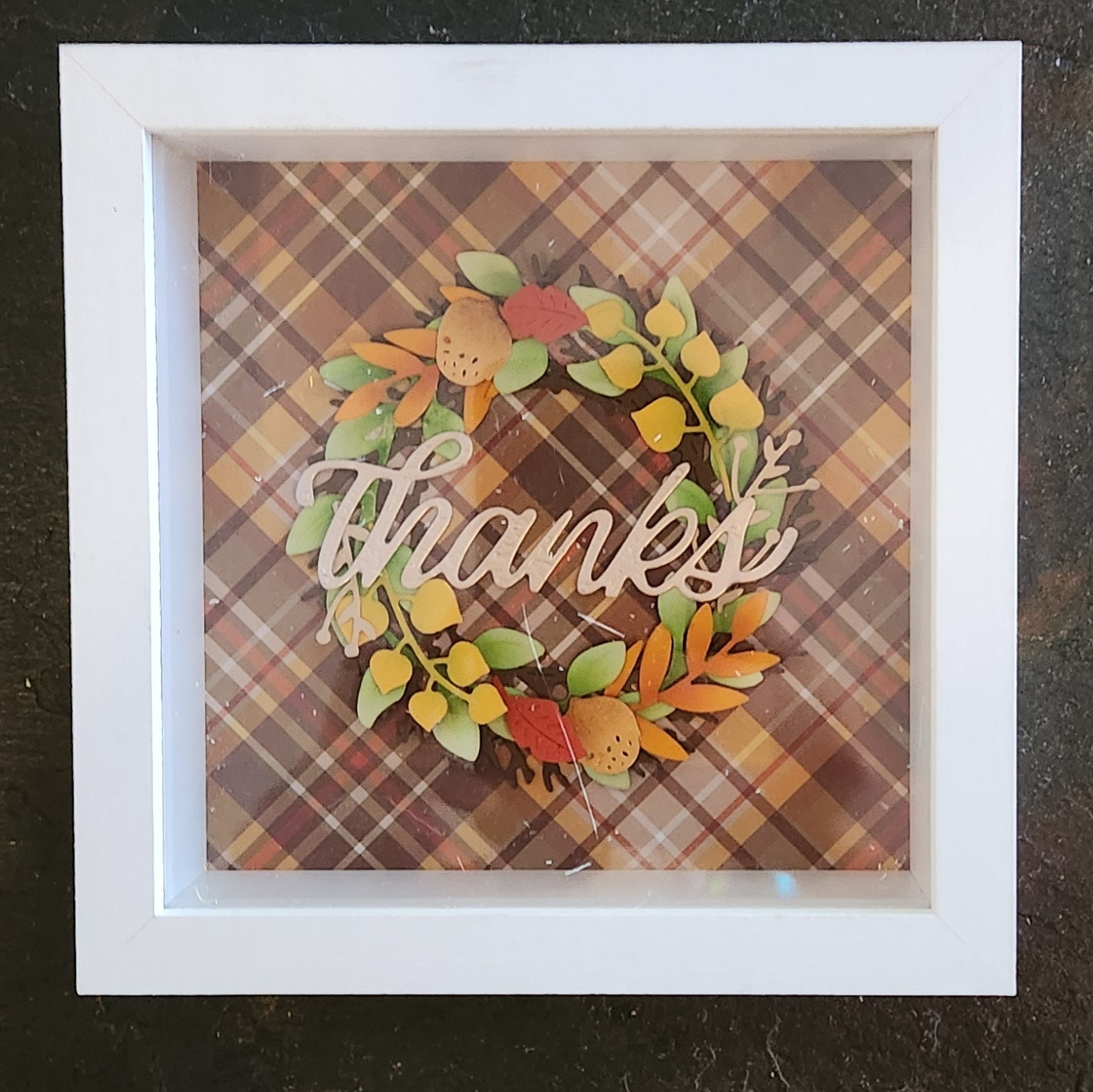 Plaid scrapbook paper in autumn colours serves as the background of this lovely diorama. The focal point is a wreath made of diecut paper twigs and leaves in various autumn colours: yellow-green, red-orange, orange, green, mustard yellow and brown. Two tiny acorns embellish the wreath. The word "thanks", diecut in a script font from hammered gold paper, lies across the wreath.