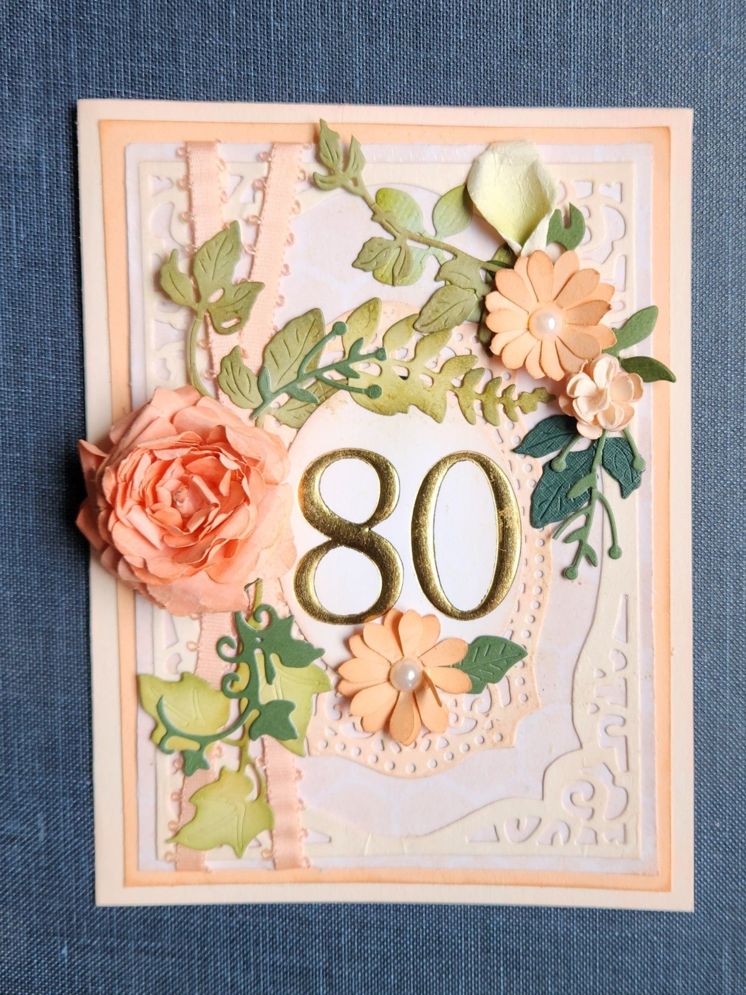 A pretty 80th birthday card in variois peach tones features 80 embpossed in gold, peach ribbon, and handsculpted paper flowers oj vines of ivy.