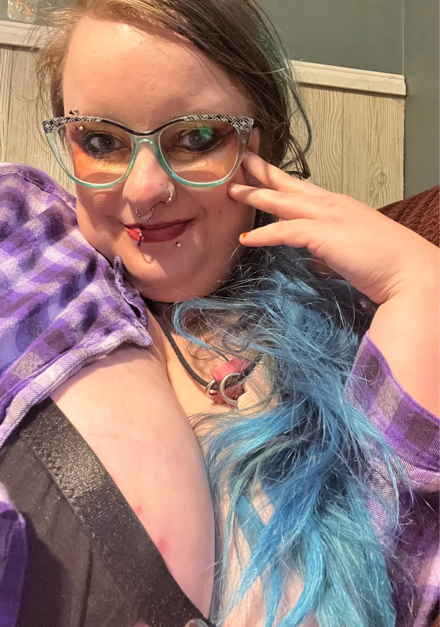 selfie of sinead with purple flannel open over black top, blue hair messy over left shoulder and left hand to cheek next to green clear cat eye glasses. fae has red and gold makeup on and is smiling softly.