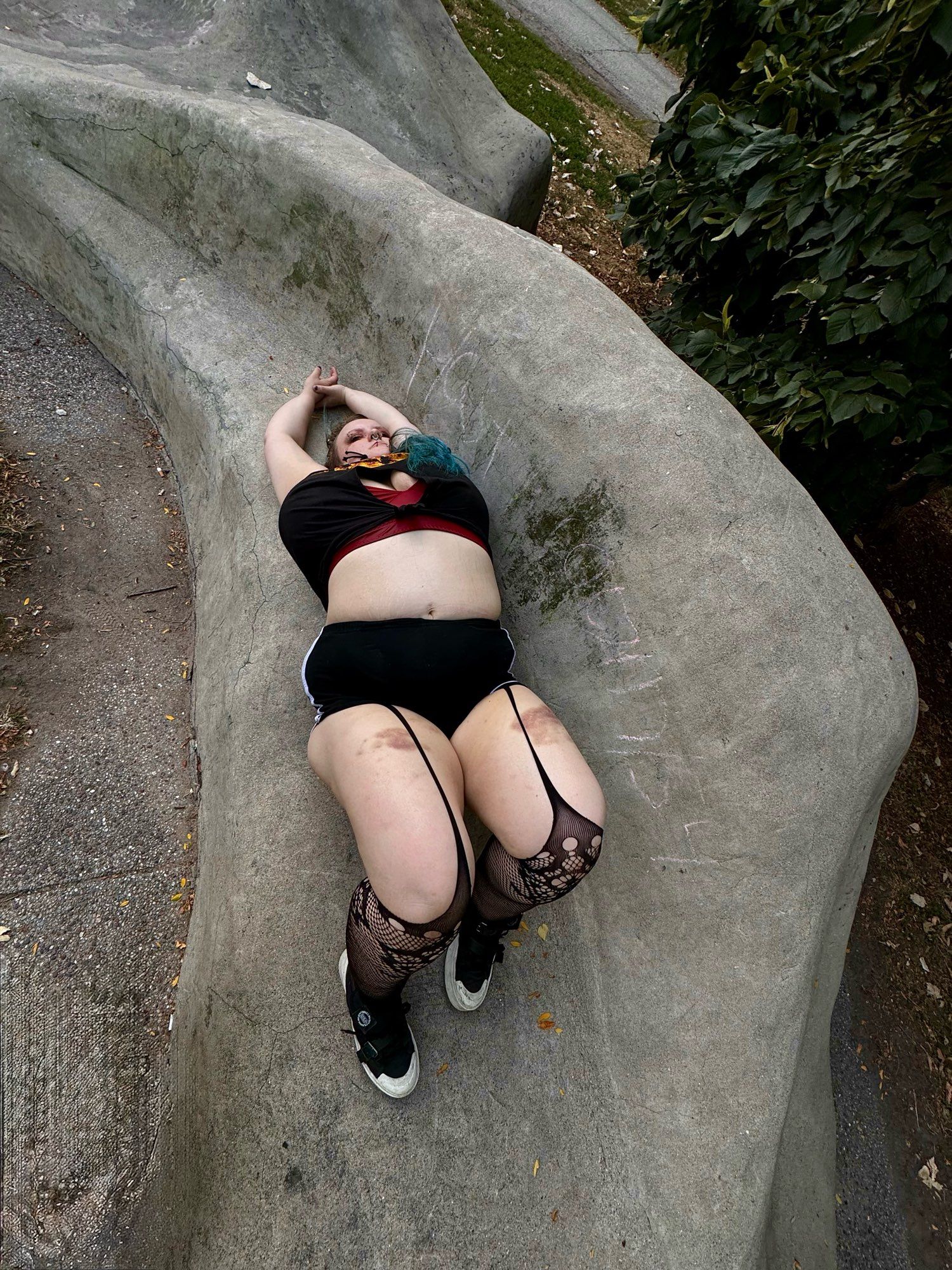 view from a distance of sinead laying on its back on a sculptural bench in black cut up tshirt showing red lingerie in underboob window, black short shorts over gartered fishnets showing bruised thighs, and black sneakers. its hands are together stretched above its head, and tummy is on full display.