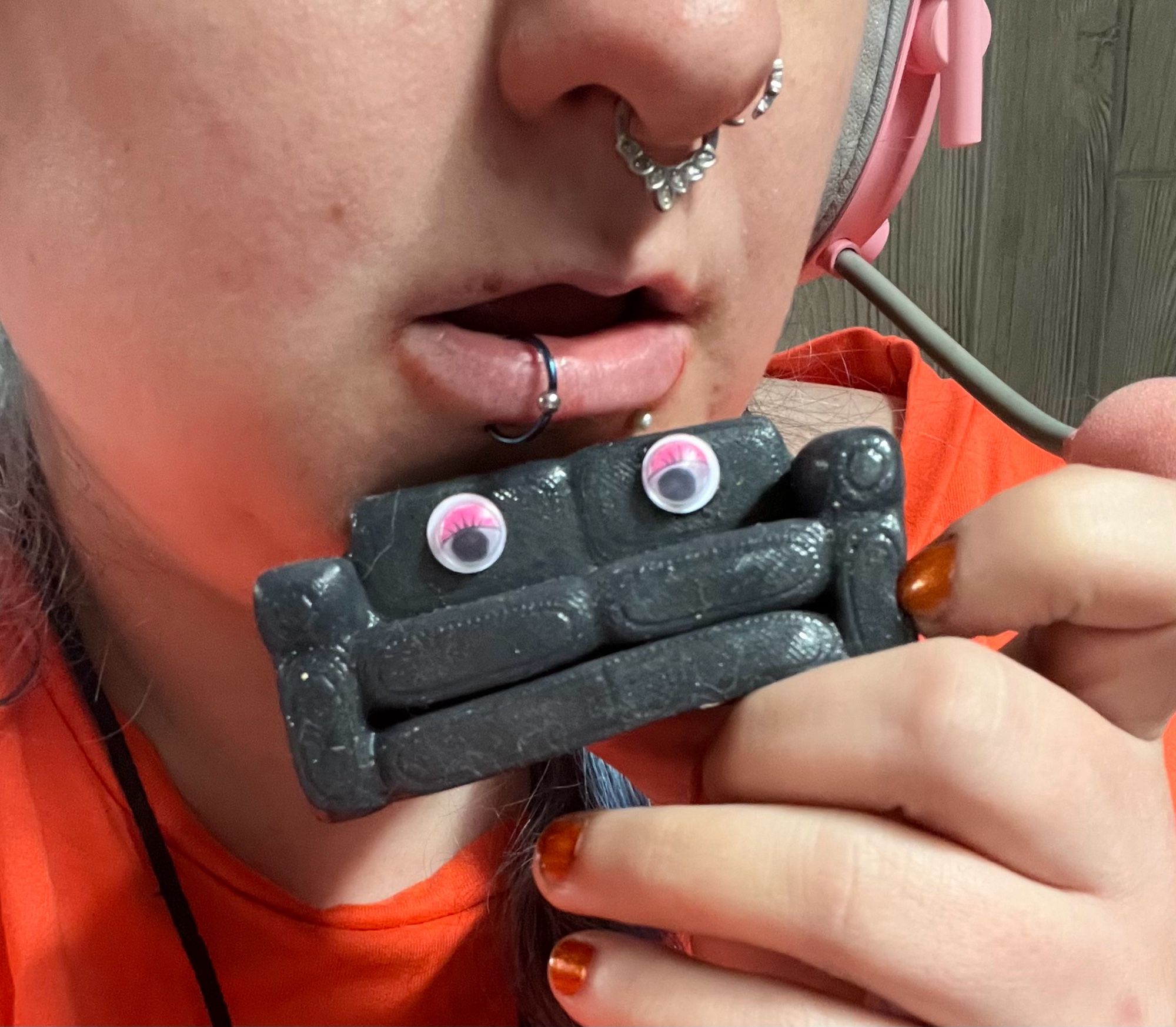 close up of sinead’s lower face with open lips above a miniature silicone couch fae is holding in orange-tipped fingers. the couch has googly eyes (the kind with pink eyeshadow and eyelashes) making a face above the fuckable cushion opening.