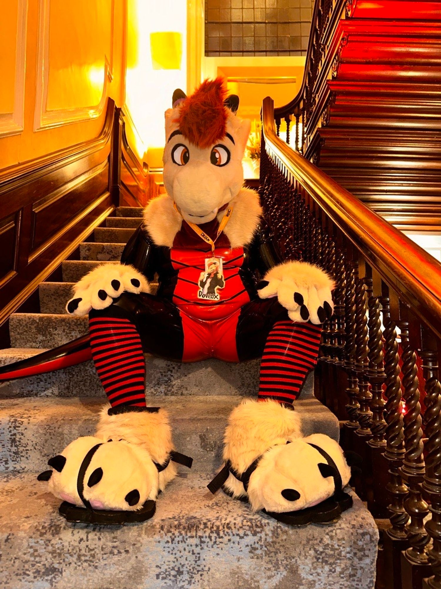 Photo of Doveux the dragon in black and red latex suit with red striped socks sat on the stairs with his cream feet and hand paws, and fursuit head.