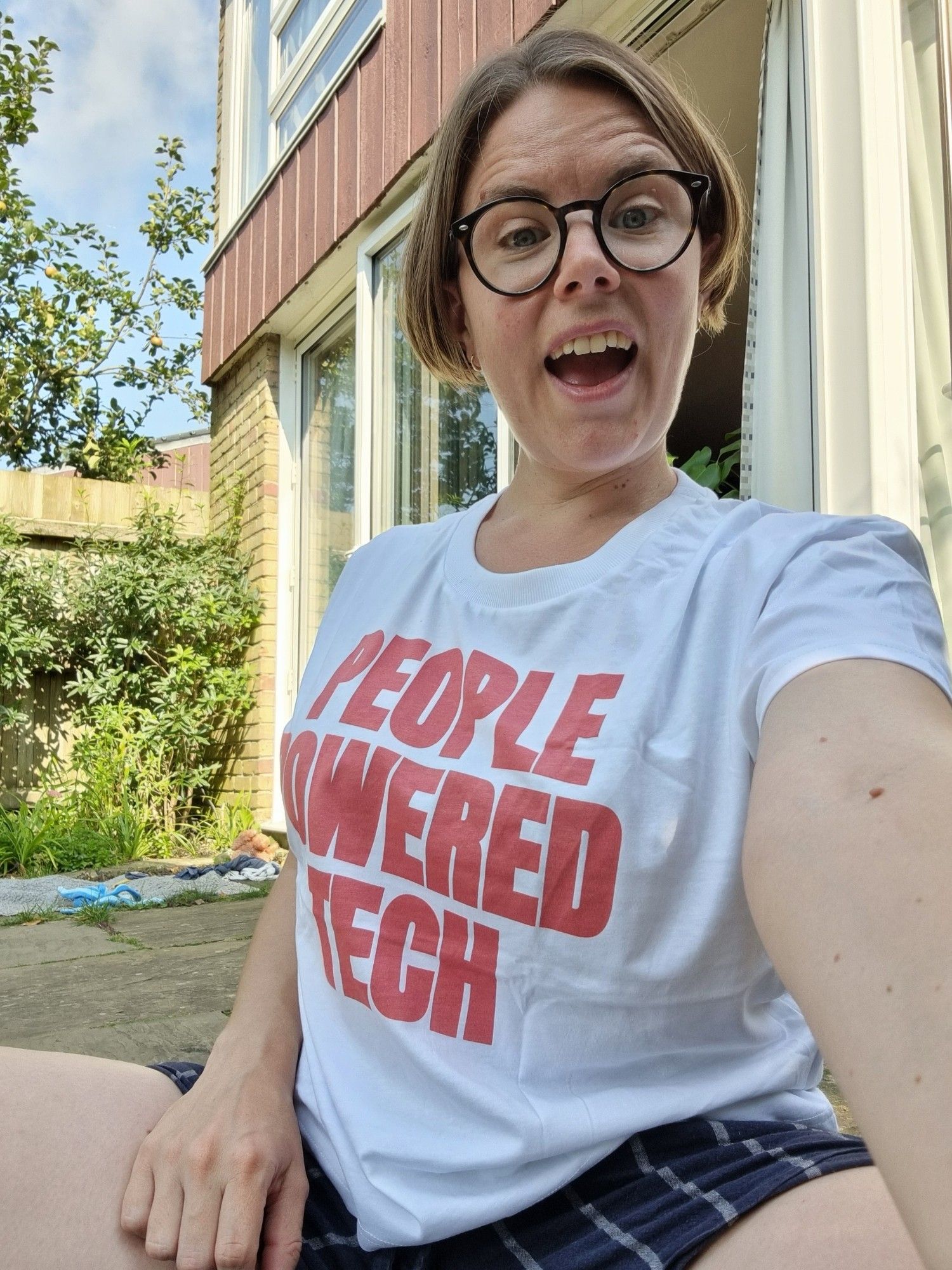 Bobi is a woman in her mid thirties sat on her patio crossed legged with a beaming smile. She is wearing a white t-shirt with the words "people powered tech" written in red across the centre.