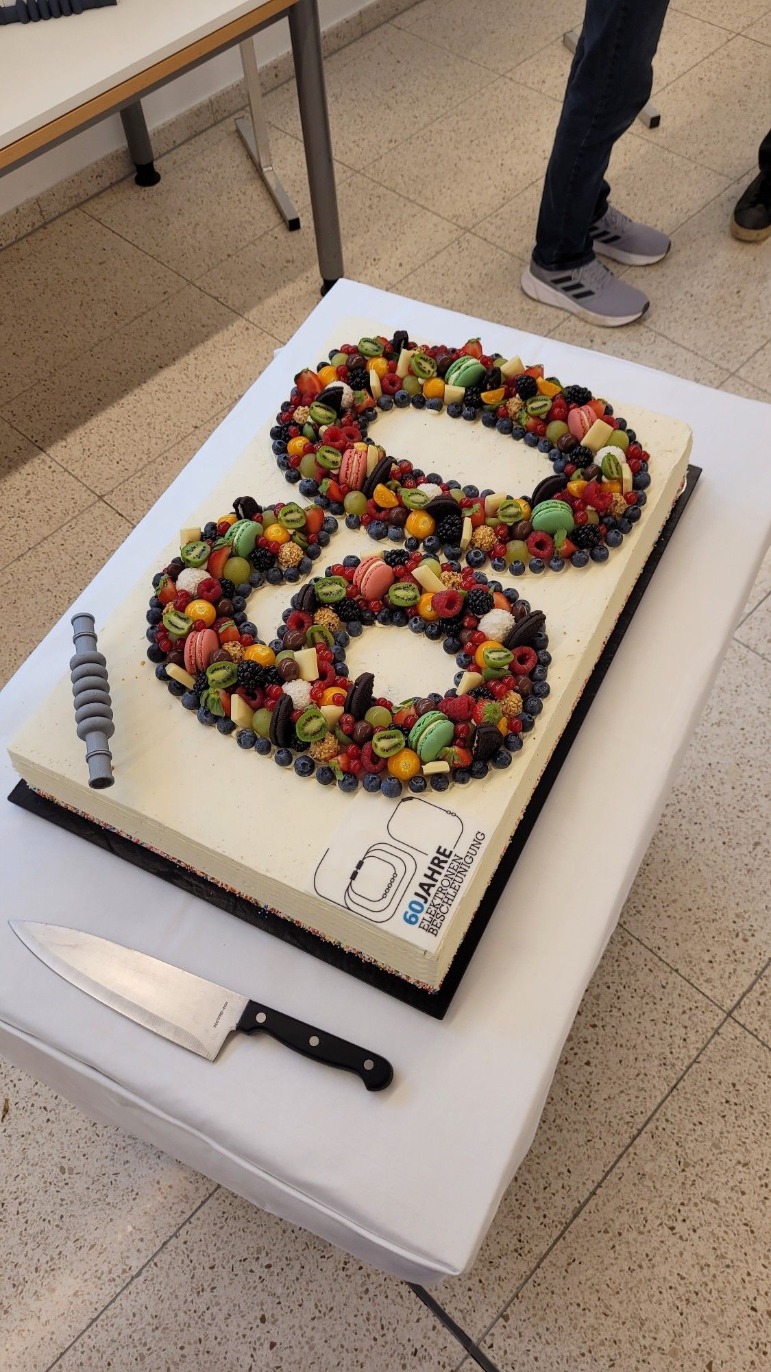 Eine große Torte mit einer bunten 60, die aus verschiedenen Obstsorten und Süßigkeiten gelegt wurde. Daneben das Logo des Jubiläums und ein 3D-gedrucktes Teil des Beschleunigers als Dekoration.