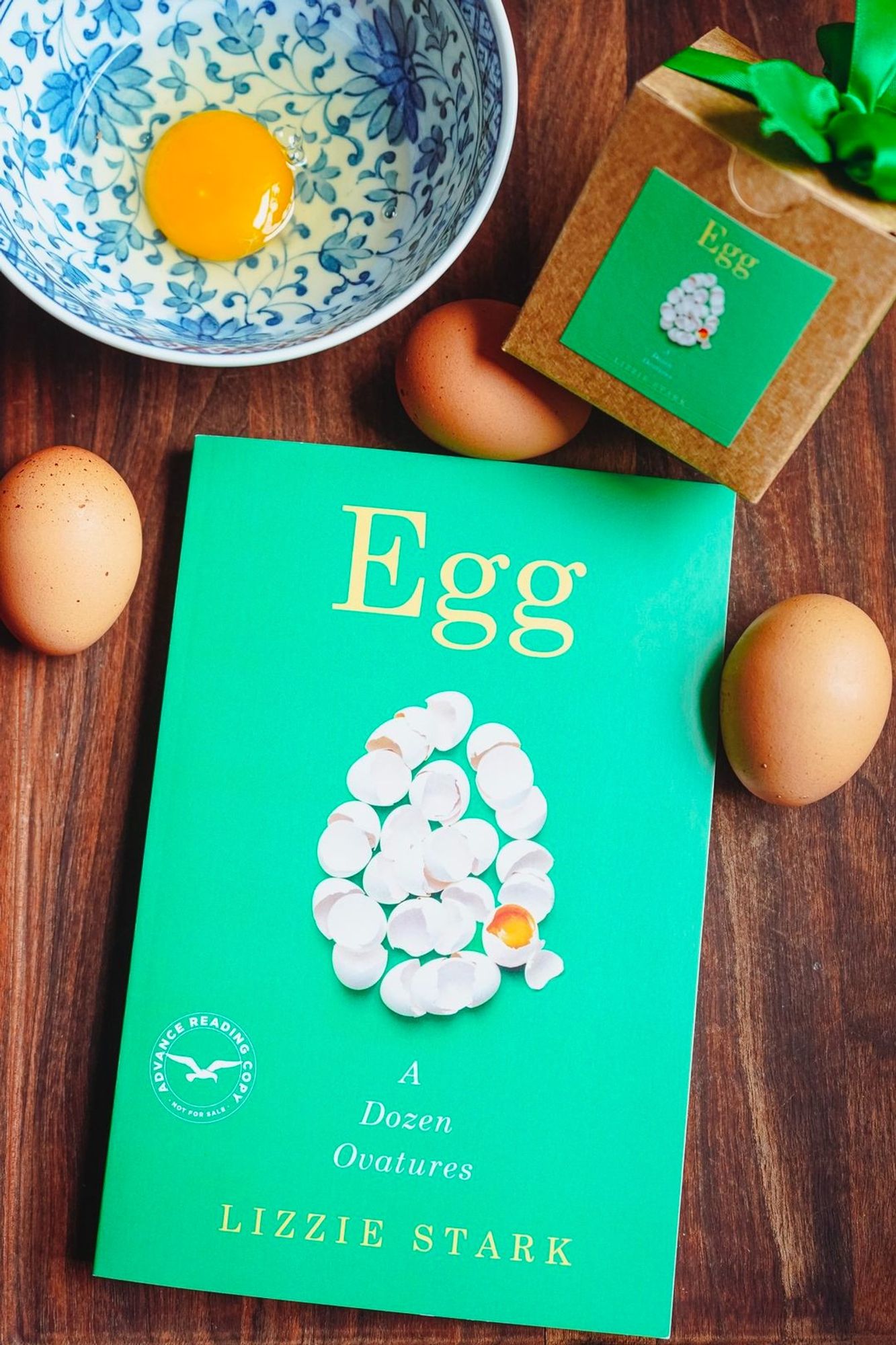 A cracked egg in a blue and white bowl sits beside several brown eggs, the bright green book EGG: A DOZEN OVATURES by Lizzie Stark, and a small brown box with an egg sticker on it.