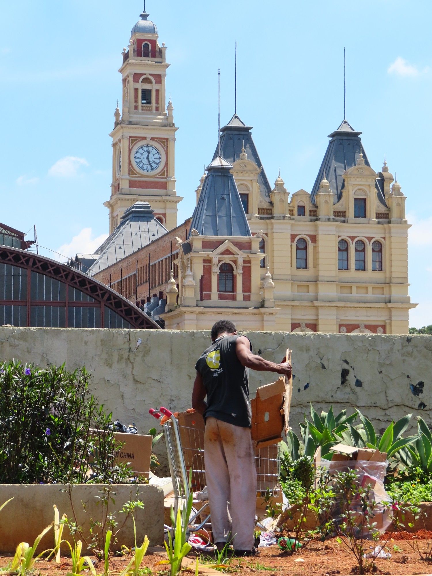 It shows São Paulo's Luz station, a famous landmark of english architecture in the background with a homeless person looking for cardboards in the trash in front of it.