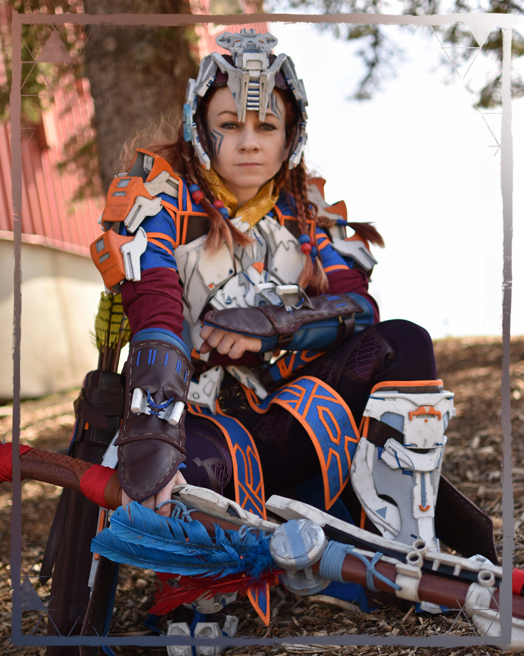 A photo of an Aloy cosplayer kneeling down in front of tree, wearing Carja armor, resting her spear down on the ground.